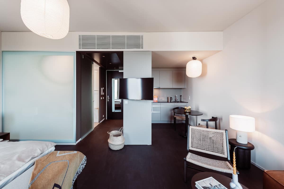 A modern hotel room with a bed, a small sitting area with a chair and table, a kitchenette, and a television.