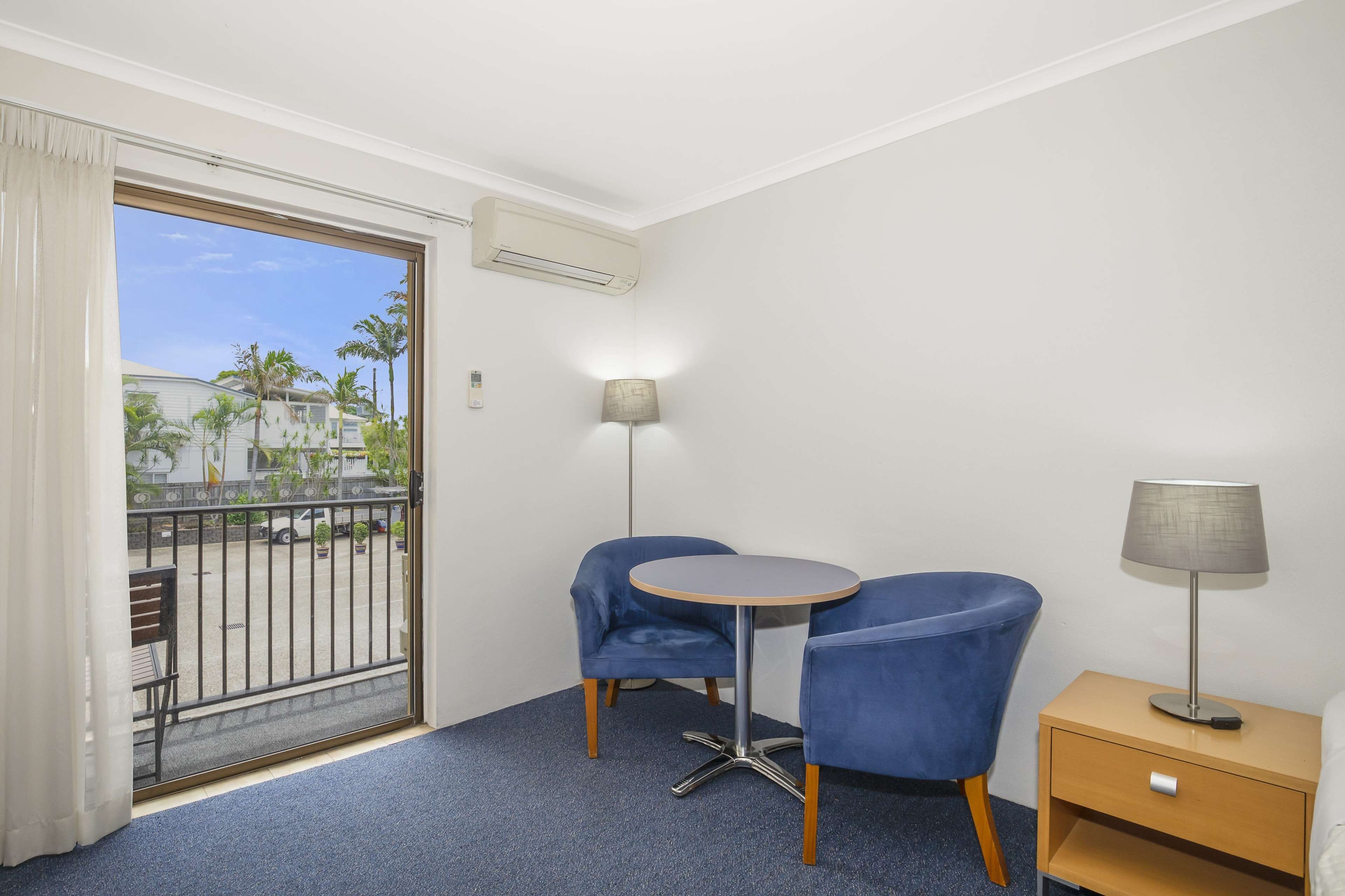 A room with a balcony, two blue chairs, a small round table, a lamp, and a nightstand.