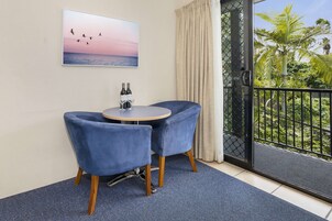 Una habitación con una mesa redonda, dos sillas azules, un cuadro de pájaros volando sobre el agua, y vistas a un balcón con plantas.