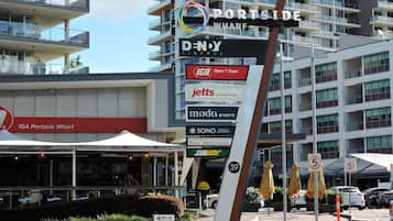 Um edifício com vários andares, com uma placa indicando 'Portside Wharf' e diversos estabelecimentos, incluindo 'IGA', 'Jetts', 'Moda', 'Sono' e 'Subway'. Há também uma placa de 'Centro de Vendas e Informações' e uma placa de 'RCQ Deliveries Portão 2'.