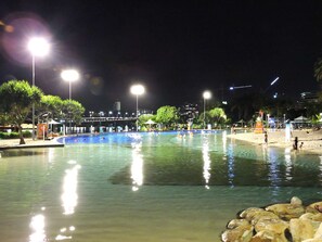 Un espace piscine nocturne avec une eau claire, entouré d’arbres et avec un pont en arrière-plan.