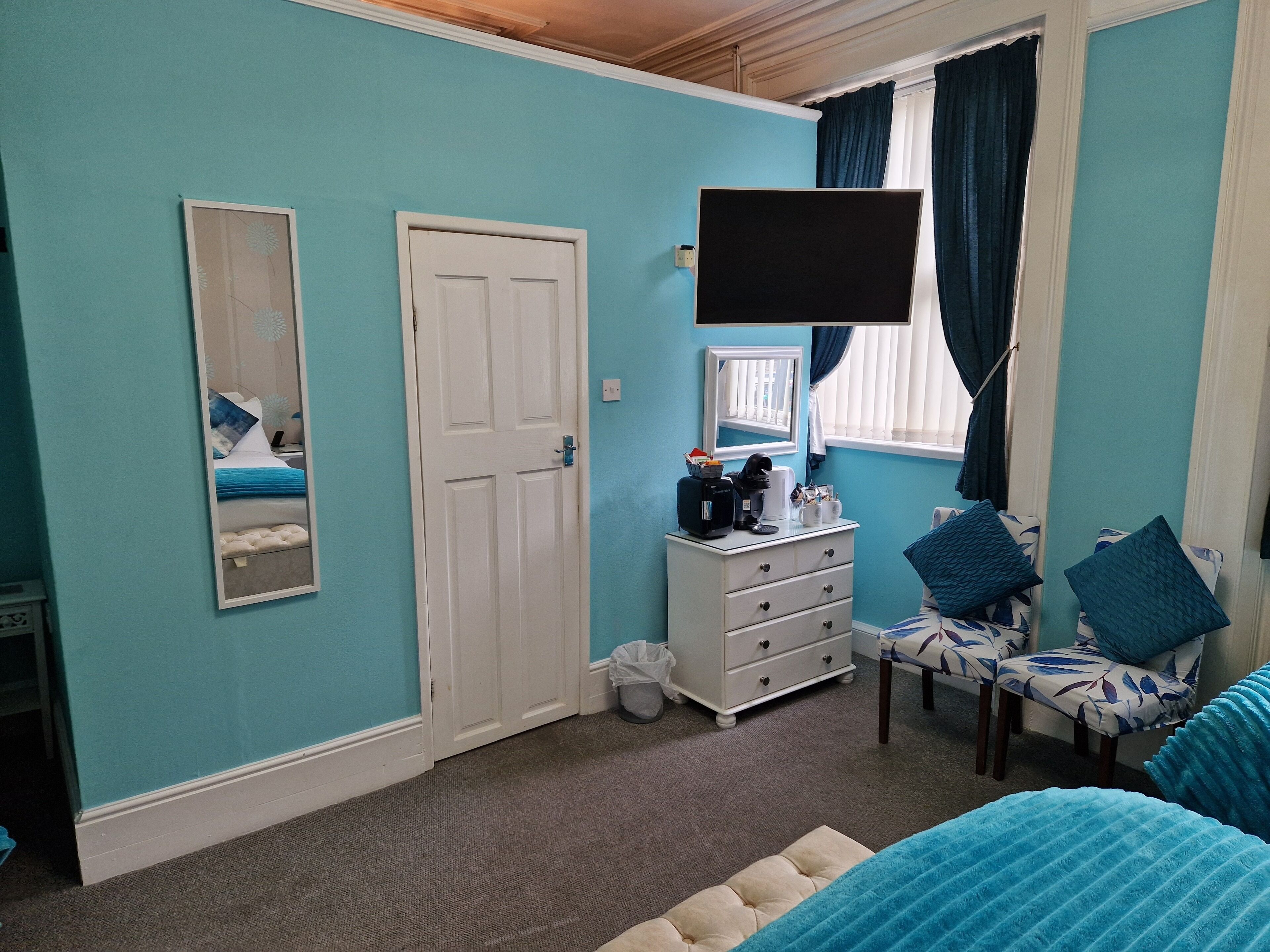A bedroom with teal walls, a white door, a dresser with drawers, a flat-screen TV mounted above a window, and a bed with blue bedding.