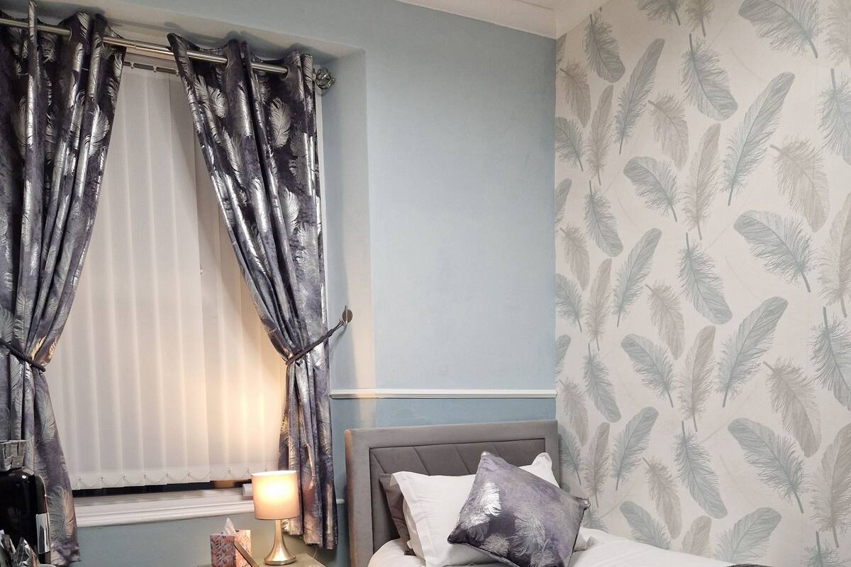A bedroom with a bed, a desk, and a chair. There is a nightstand with a lamp and a tissue box. The room has a window with curtains and a wall with a feather pattern.