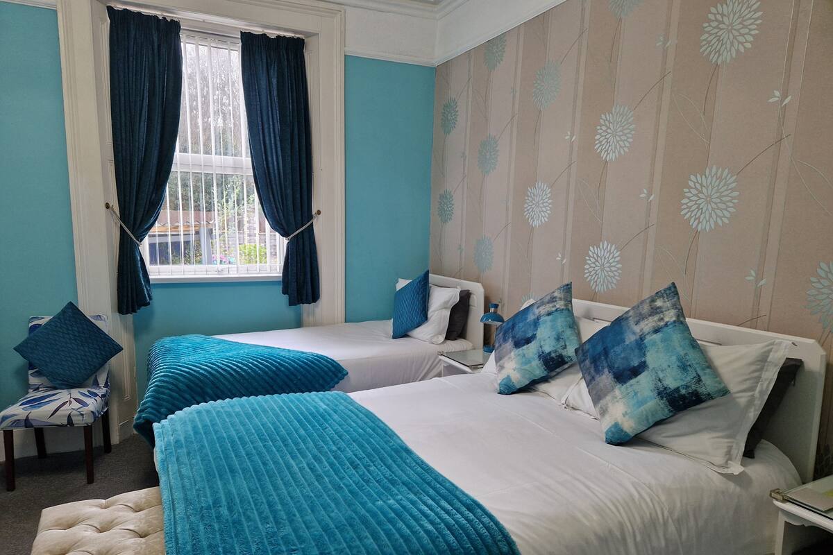 A bedroom with two beds, a window with curtains, a chair, and a wall with a floral pattern.