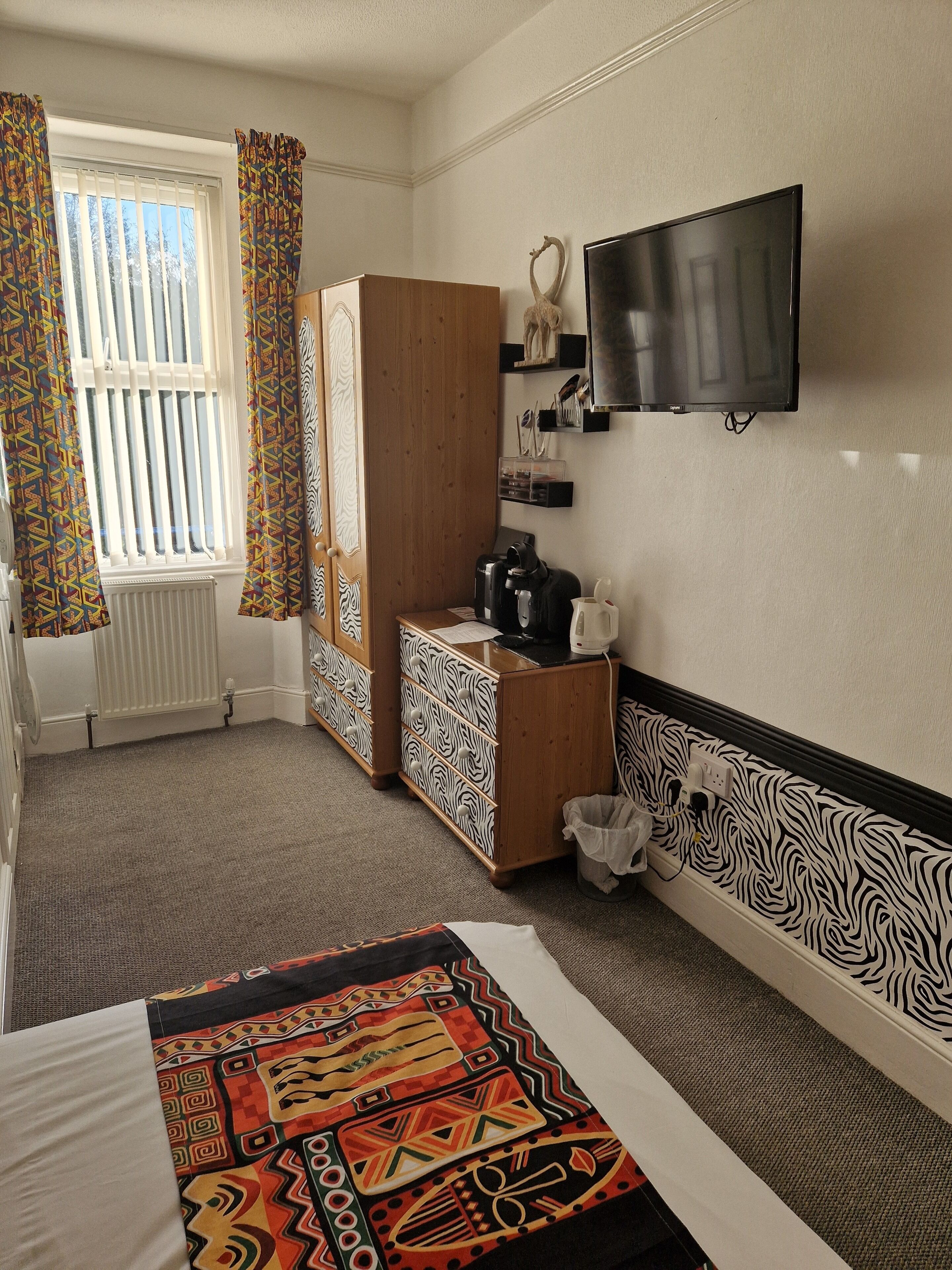 Une chambre d’hôtel avec un lit, un bureau doté d’une machine à café, une télévision, un radiateur et des rideaux à motifs.