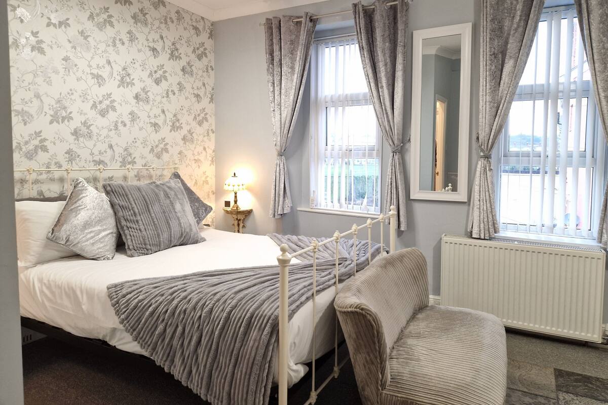 A bedroom with a bed, bedside table, lamp, chair, window, and radiator.