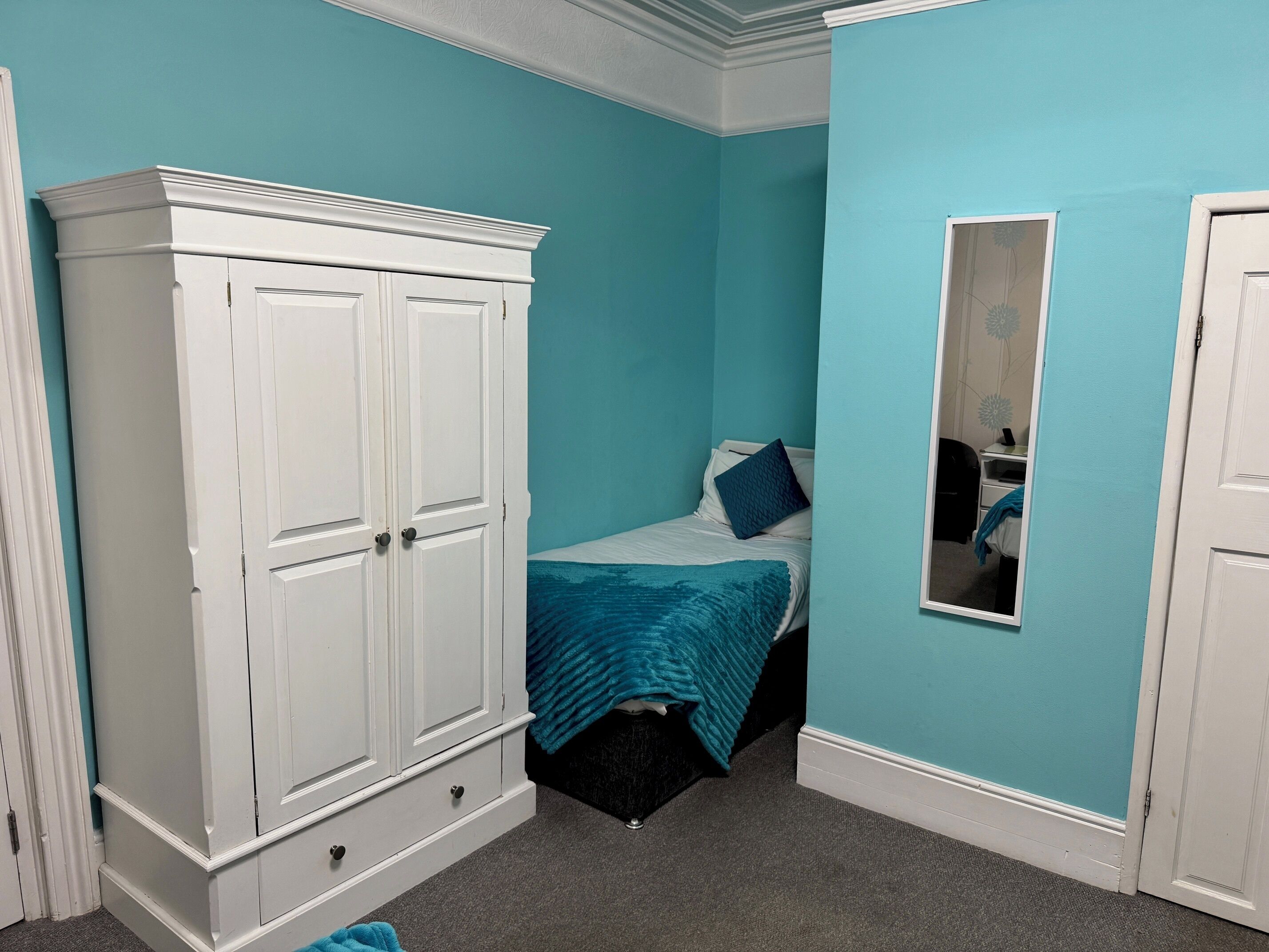 Un dormitorio con paredes color turquesa, un armario blanco, una cama con manta azul y un espejo en la pared.