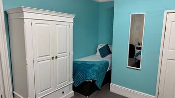 Un dormitorio con paredes color turquesa, un armario blanco, una cama con manta azul y un espejo en la pared.