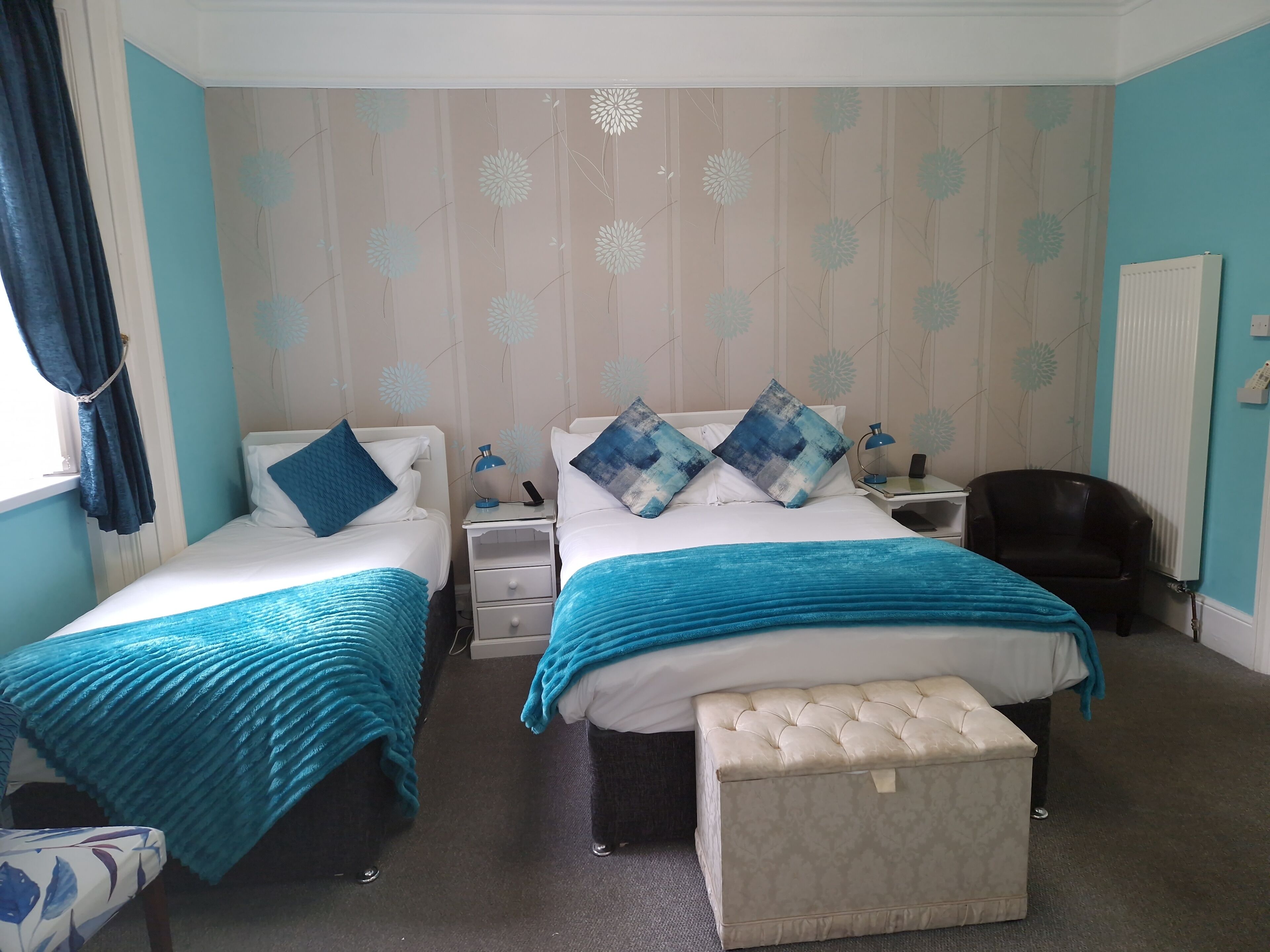 A hotel room with two beds, a small table, a chair, a radiator, and a window with curtains.