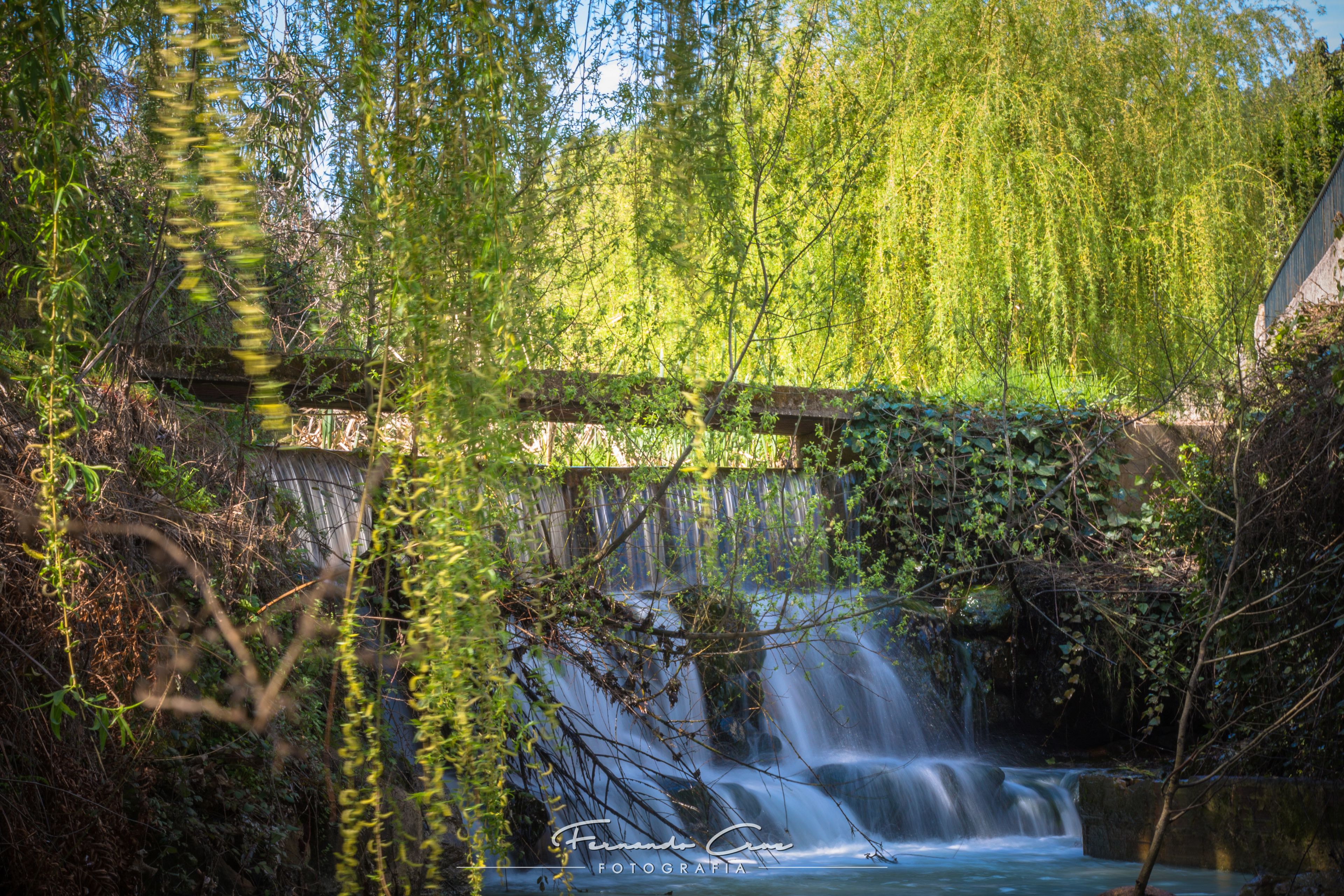 A waterfall cascading over rocks, surrounded by lush greenery and a wooden bridge.