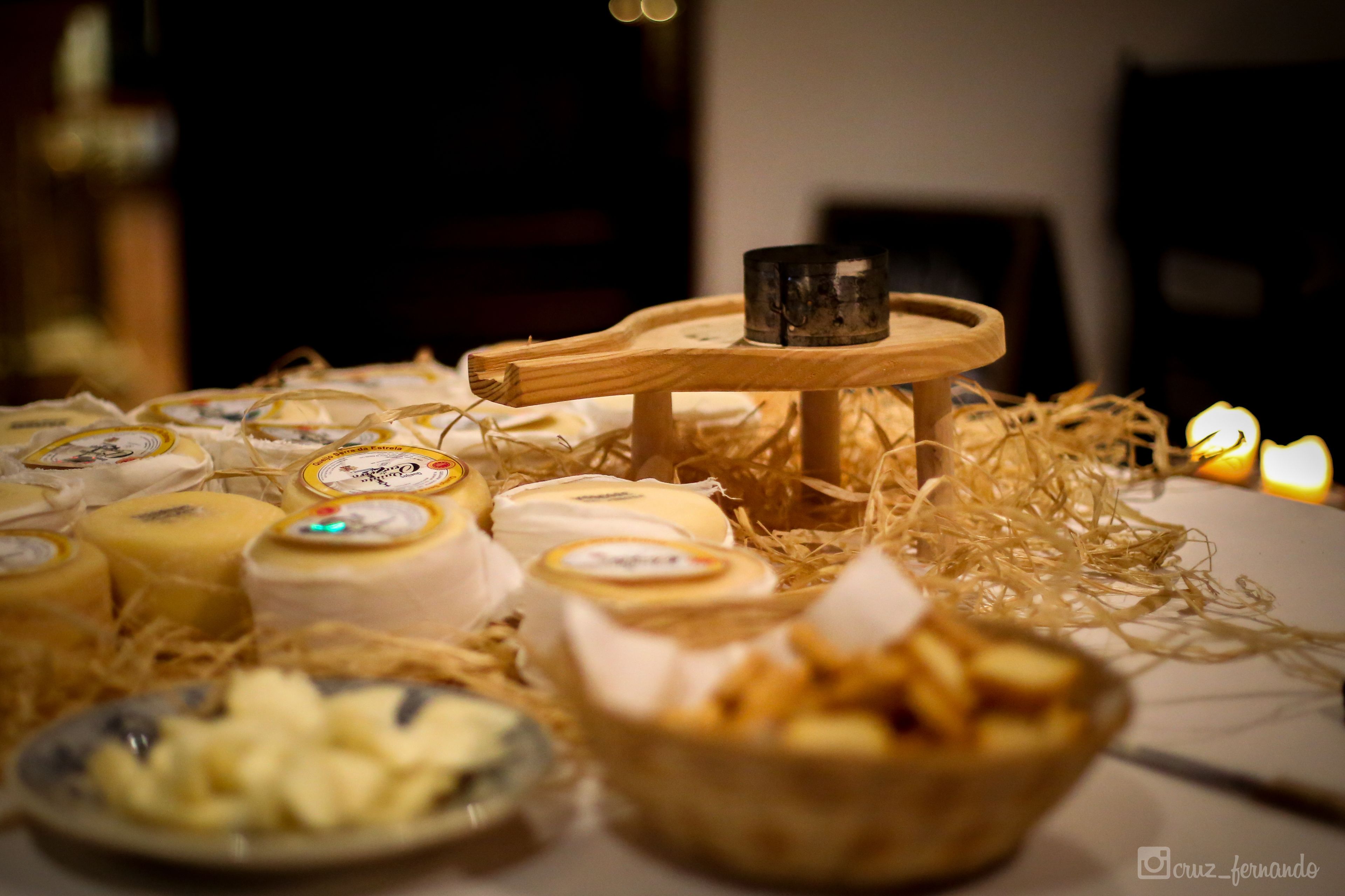 A variety of cheeses on a table with a wooden stand in the center, surrounded by shredded paper and candles.