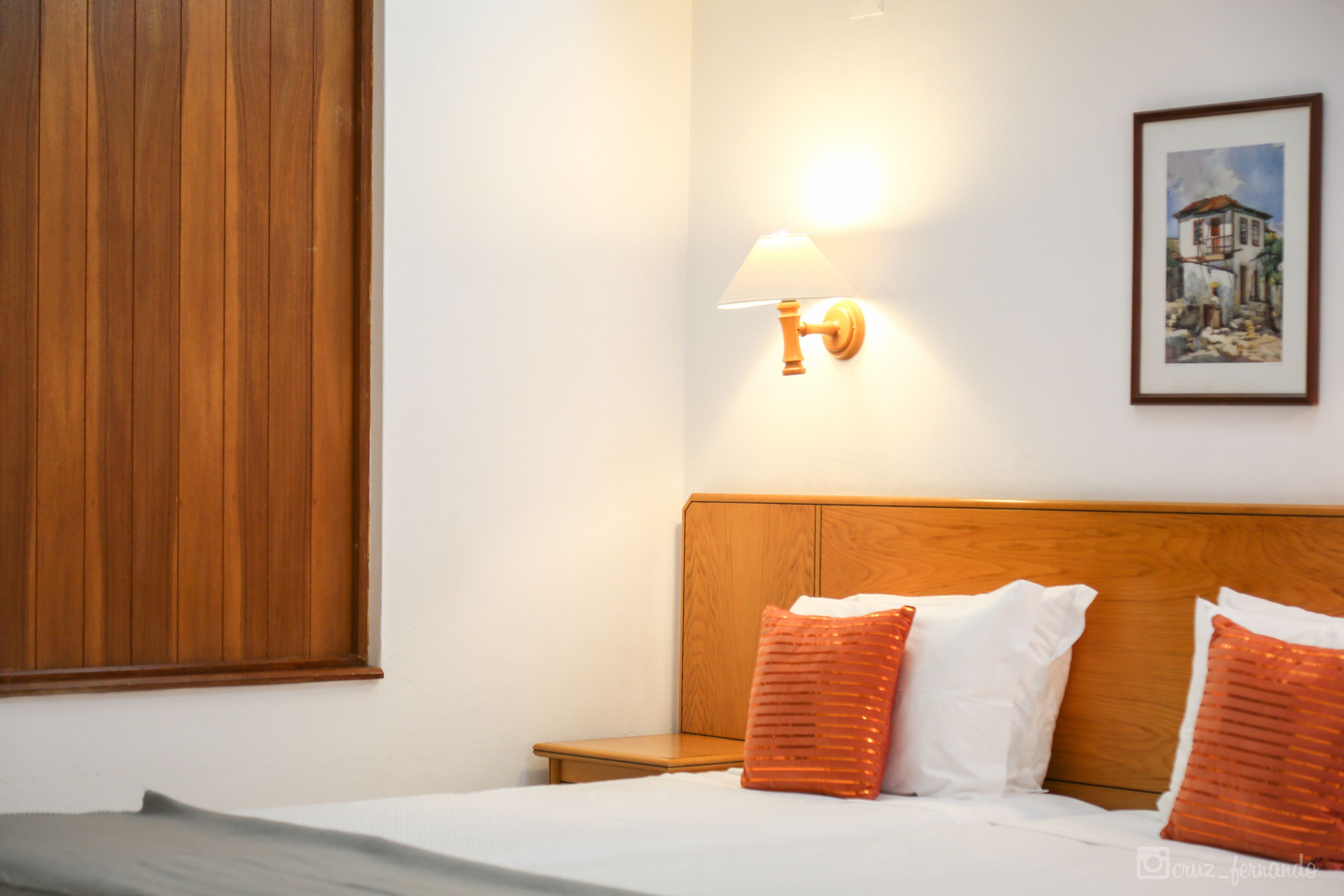A hotel room with a wooden headboard, a bedside table, a lamp, and a framed picture on the wall.