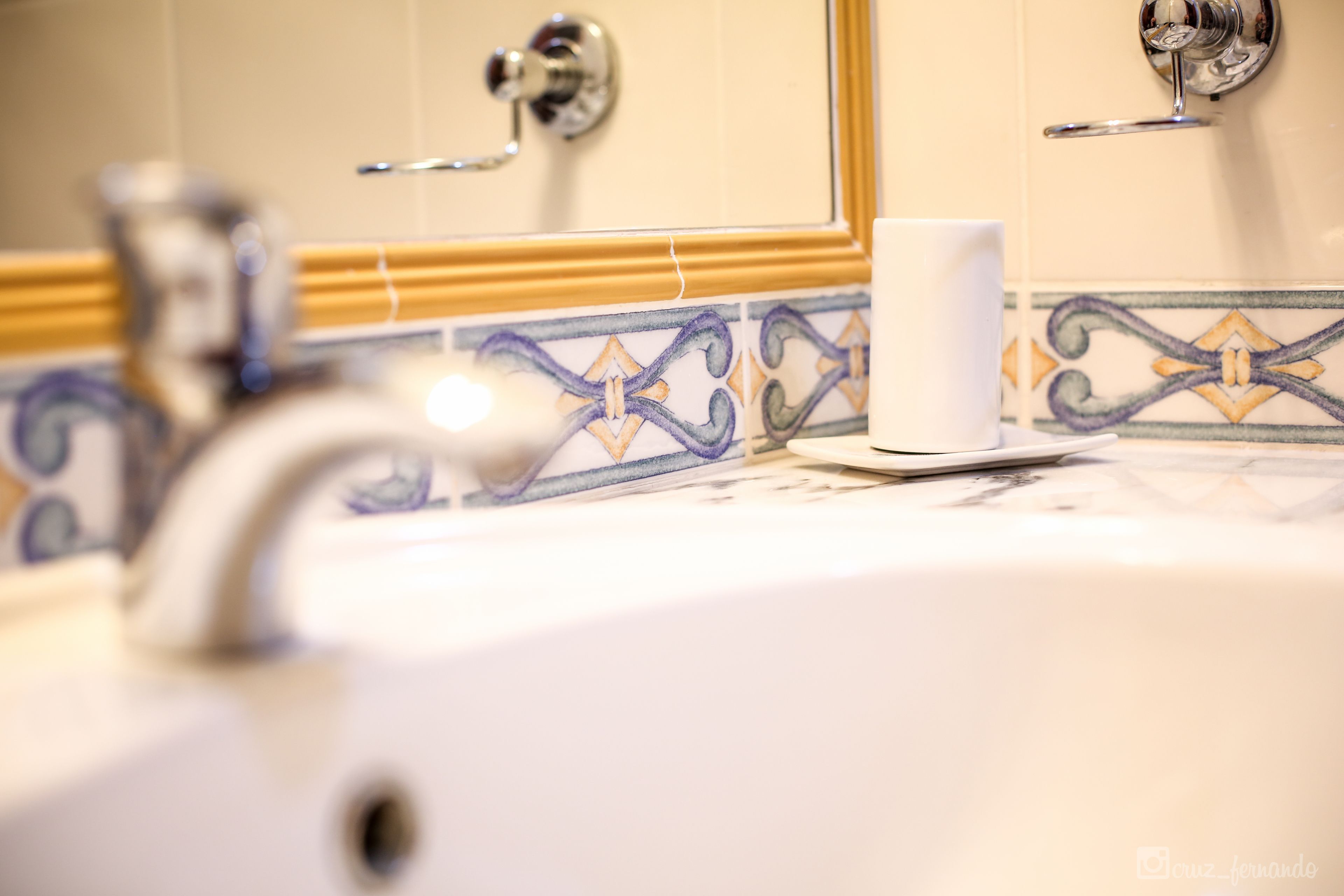 Un lavamanos con una taza blanca, un espejo y azulejos decorativos.