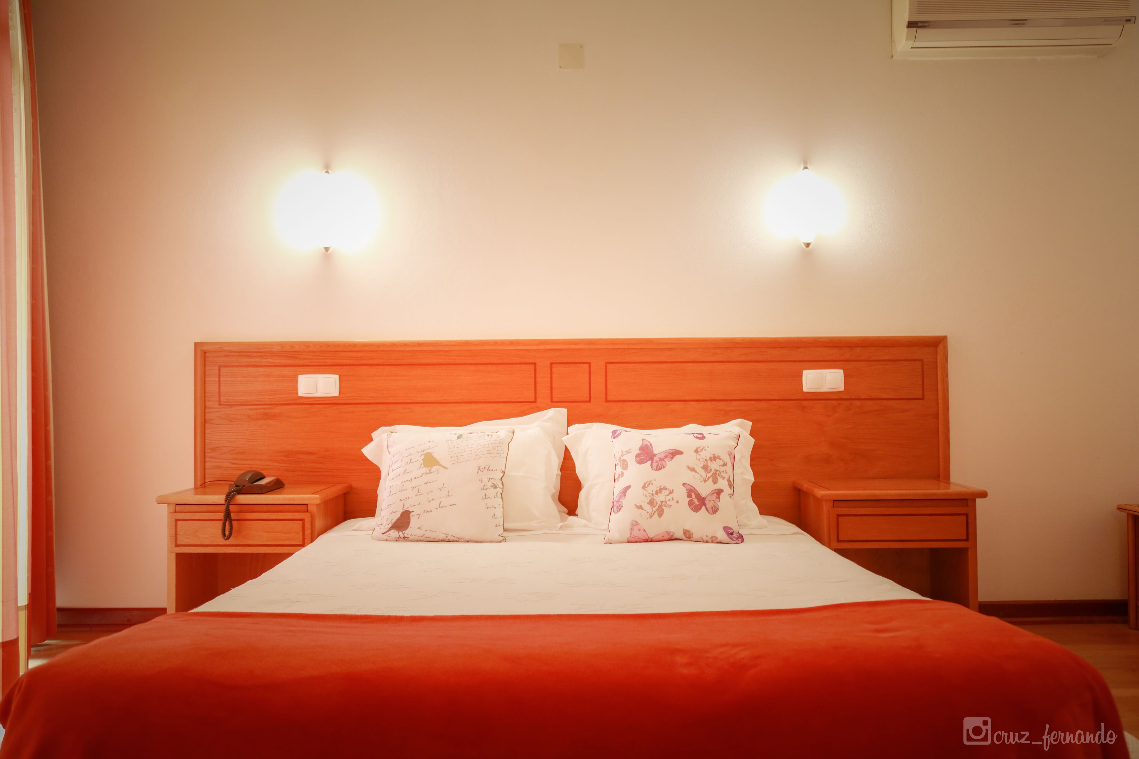 A neatly made bed with a red bedspread, two bedside tables with lamps, and a wooden headboard.