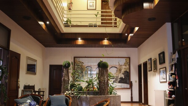 A spacious lobby with a high ceiling, wooden flooring, and a central seating area with chairs and a red rug. There are potted plants and a large painting on the wall.