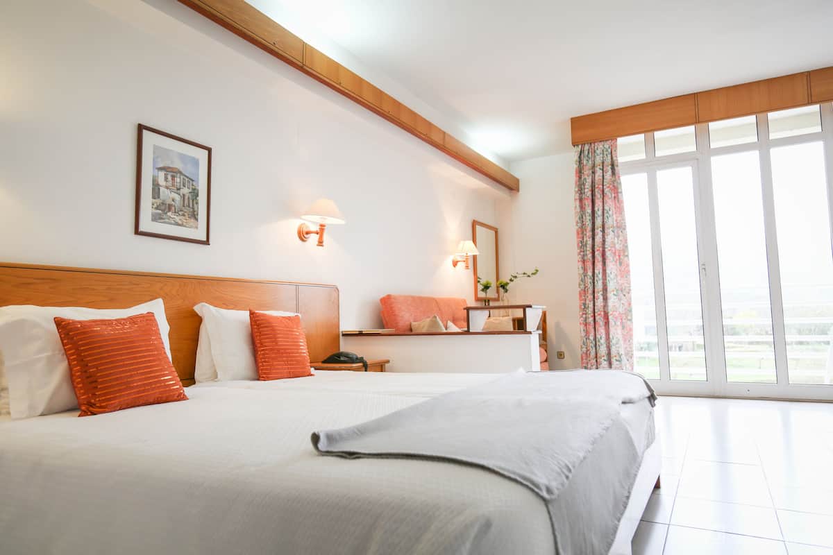 Ein Hotelzimmer mit zwei Betten, einem Holzkopfende, einem gerahmten Bild an der Wand und einem großen Fenster mit Vorhängen.