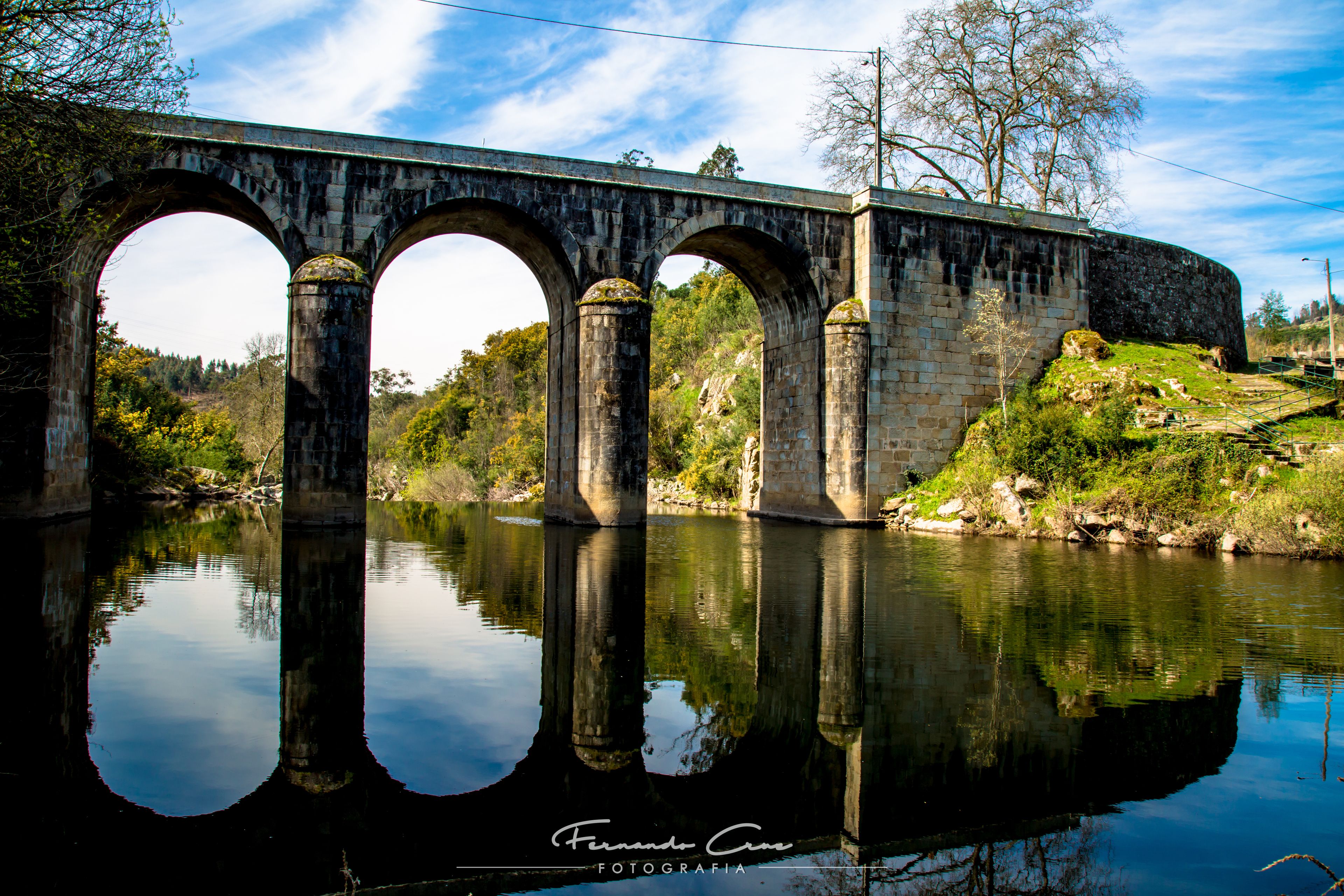 Eine Steinbrücke mit drei Bögen über einem ruhigen Fluss, umgeben von Grünflächen und Bäumen.