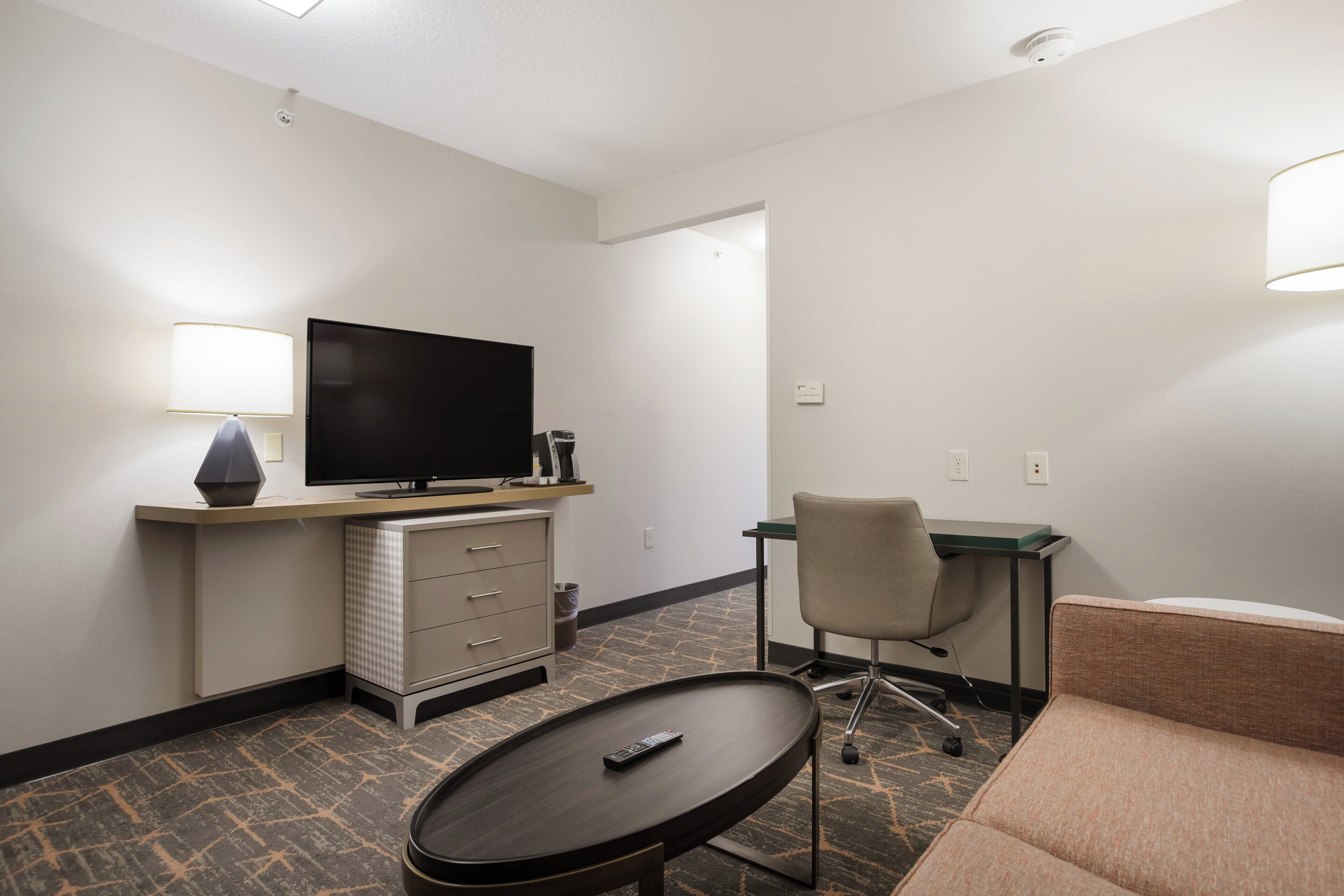A hotel room with a flat-screen TV, a desk, a chair, a sofa, a small table, and a remote control.