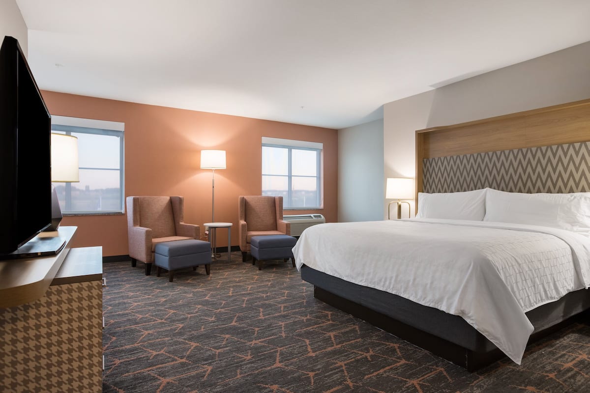 A hotel room with a large bed, two armchairs, a desk with a TV, and two windows.