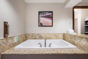 A bathroom with a large white bathtub, a handrail, a picture of a pink car on the wall, and a tiled backsplash.