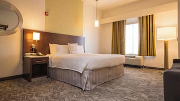 A hotel room with a large bed, a bedside table with a lamp, a round mirror, a window with curtains, and a carpeted floor.