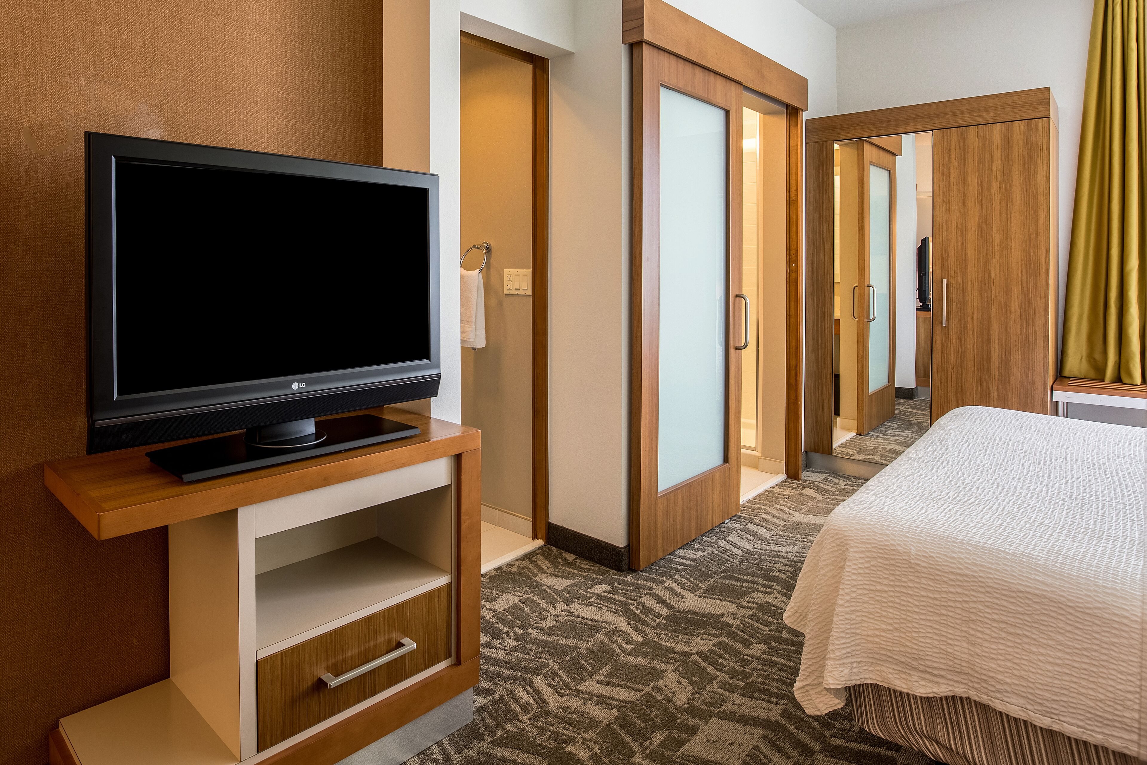 Hotelkamer met flatscreen televisie op een houten standaard, een bed met witte dekbedovertrek, een houten kledingkast en een vloerbedekking.