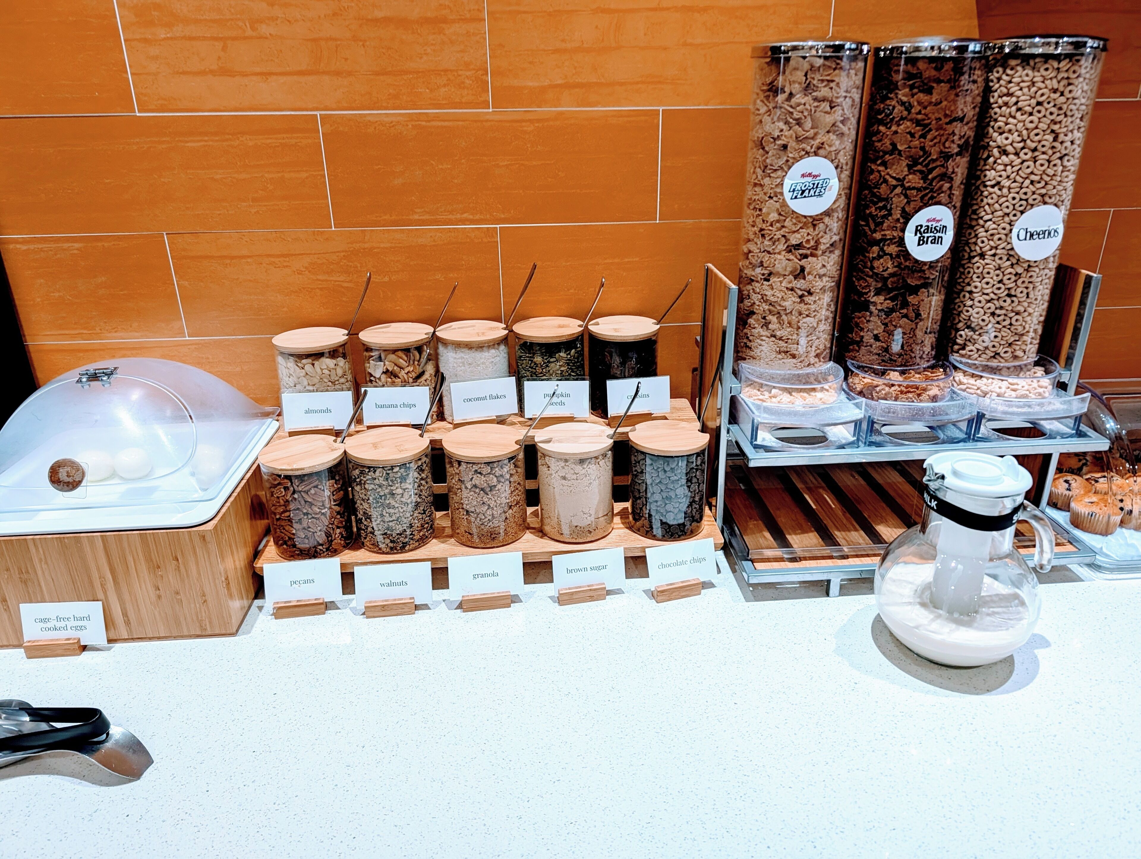 Un bufé de desayuno con recipientes etiquetados que contienen diversos alimentos como almendras, plátanos y granola.