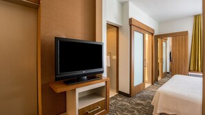 Habitación de hotel con televisión de pantalla plana sobre un soporte de madera, cama con cubrecama blanco y alfombra estampada.