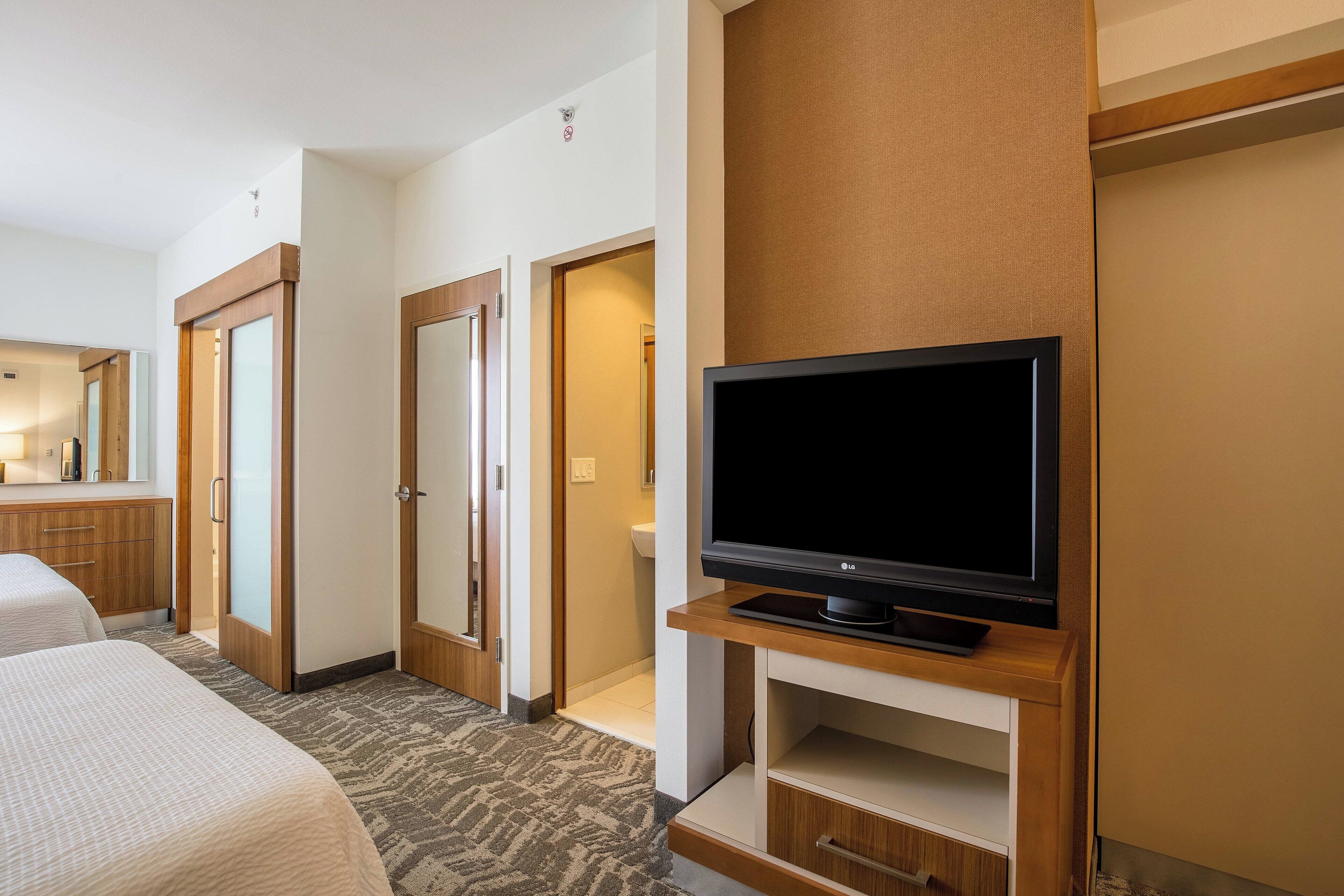 A hotel room with a television on a wooden stand, a bed, a bathroom, and a hallway leading to another room.