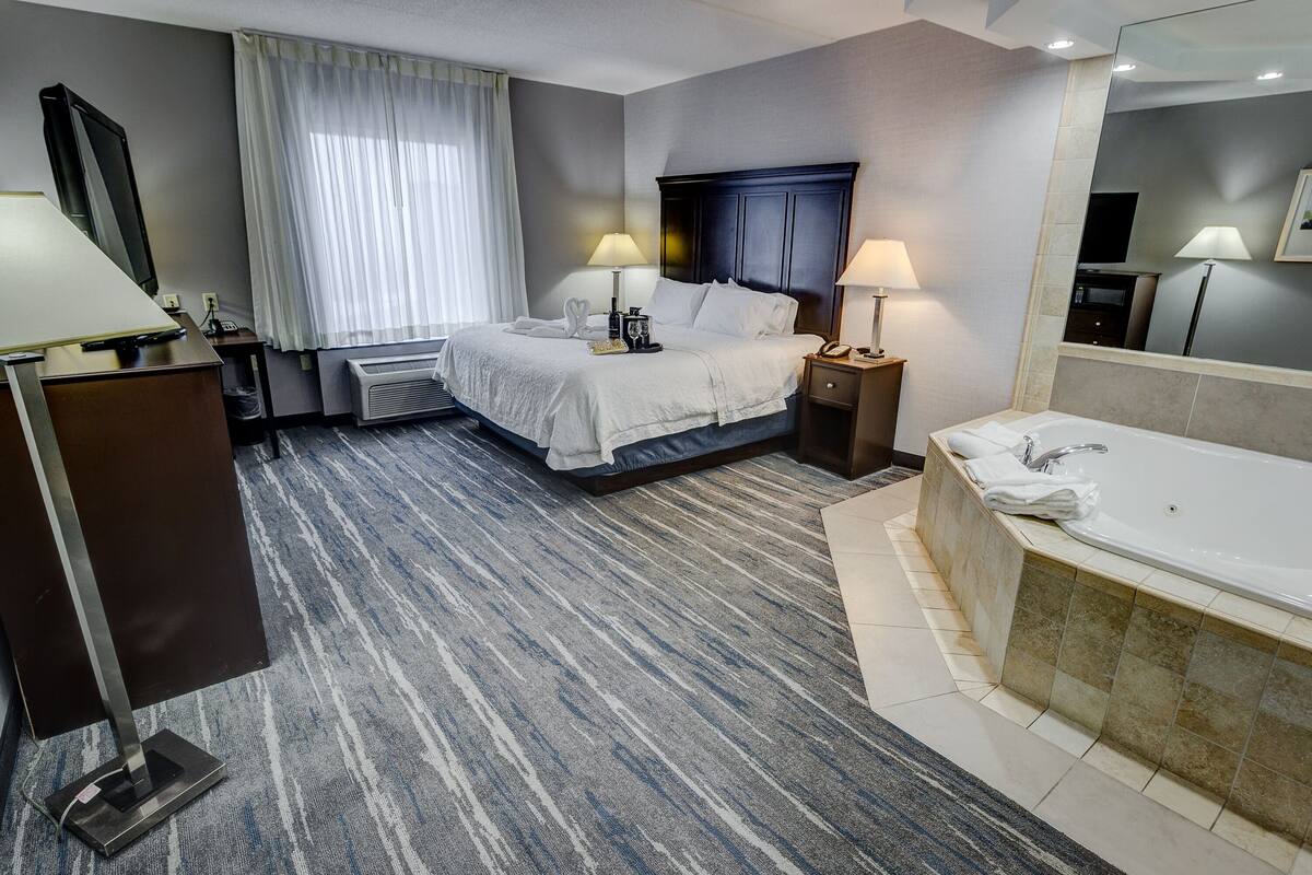 A hotel room with a bed, bedside lamps, a TV, a desk, a desk lamp, a large mirror, a bathtub, and a window with curtains.
