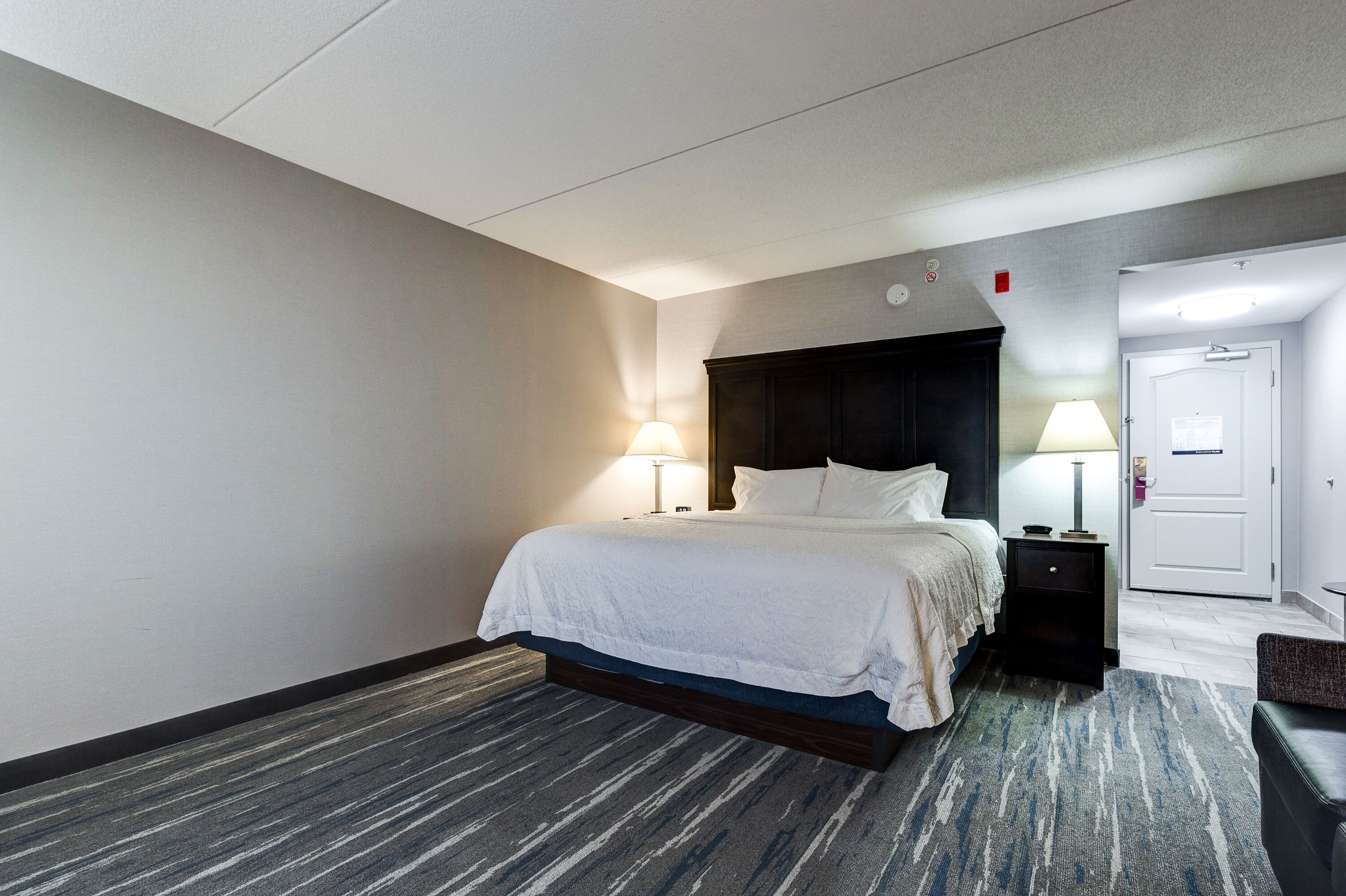 Quarto de hotel com uma cama grande, duas luminárias de cabeceira, uma cabeceira escura, um criado-mudo pequeno e uma porta para outro espaço.