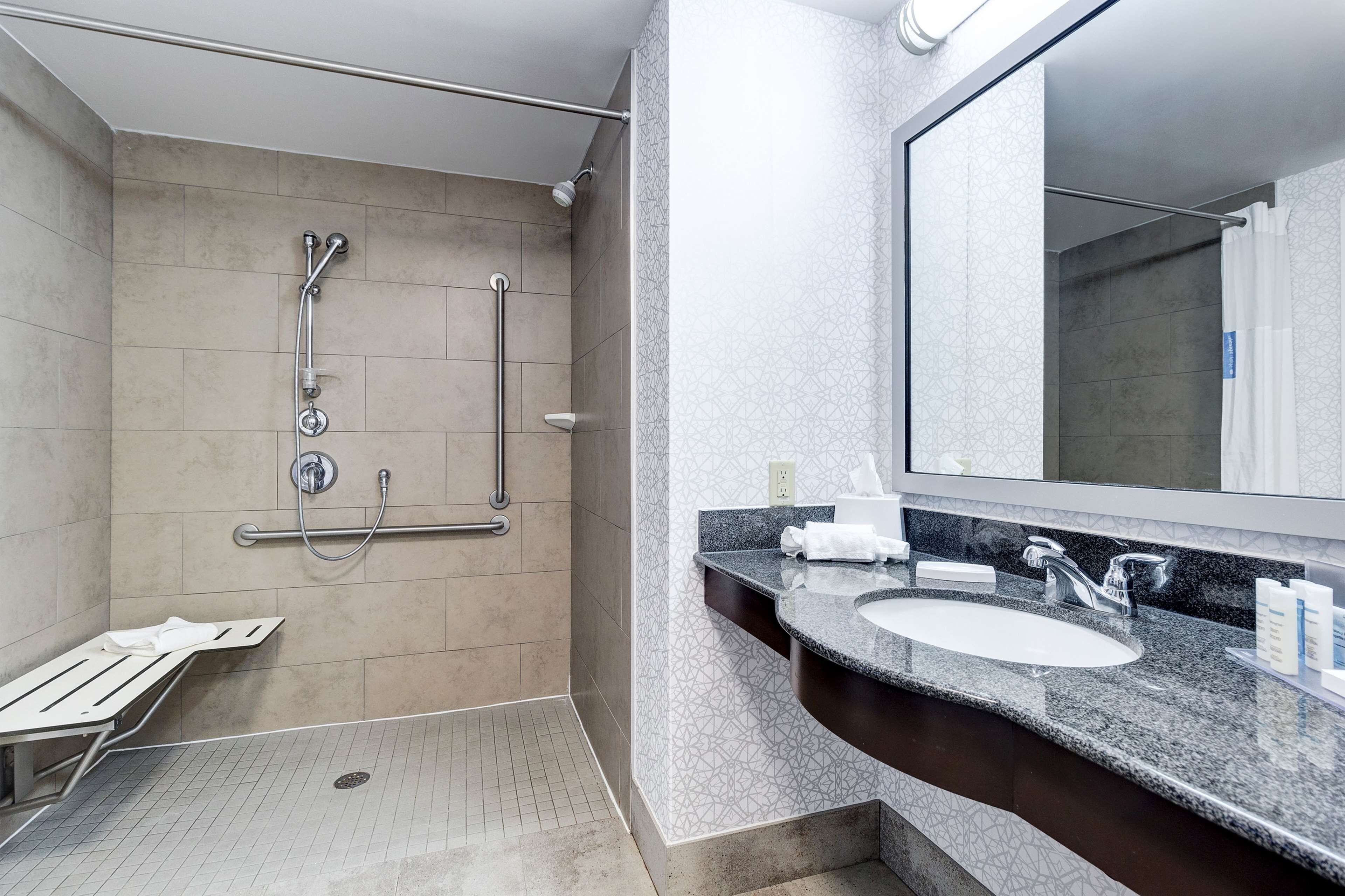 Une salle de bain moderne dotée d’une douche à l’italienne, de barres d’appui, d’un banc, d’un lavabo et d’un grand miroir.