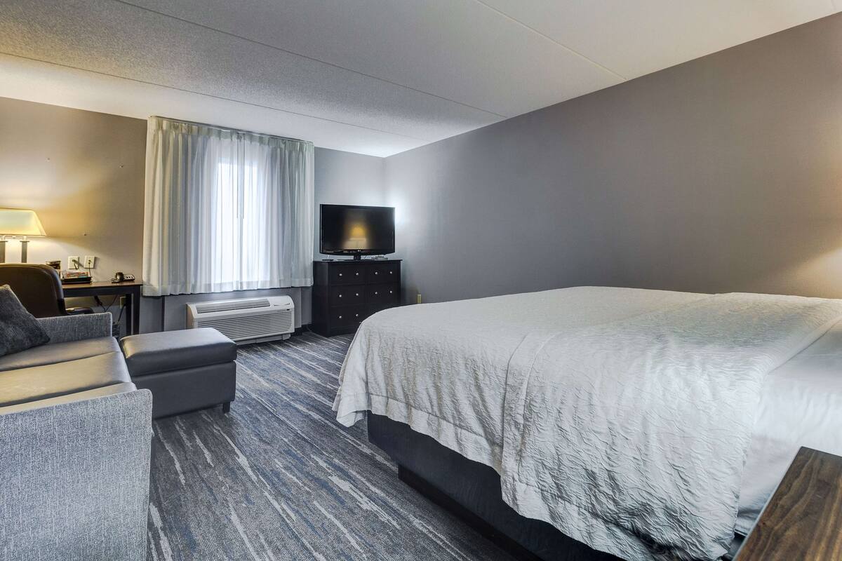 A hotel room with a large bed, a TV on a stand, a desk with a lamp, and a window with curtains.