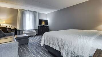 A hotel room with a large bed, a TV on a stand, a desk with a lamp, and a window with curtains.