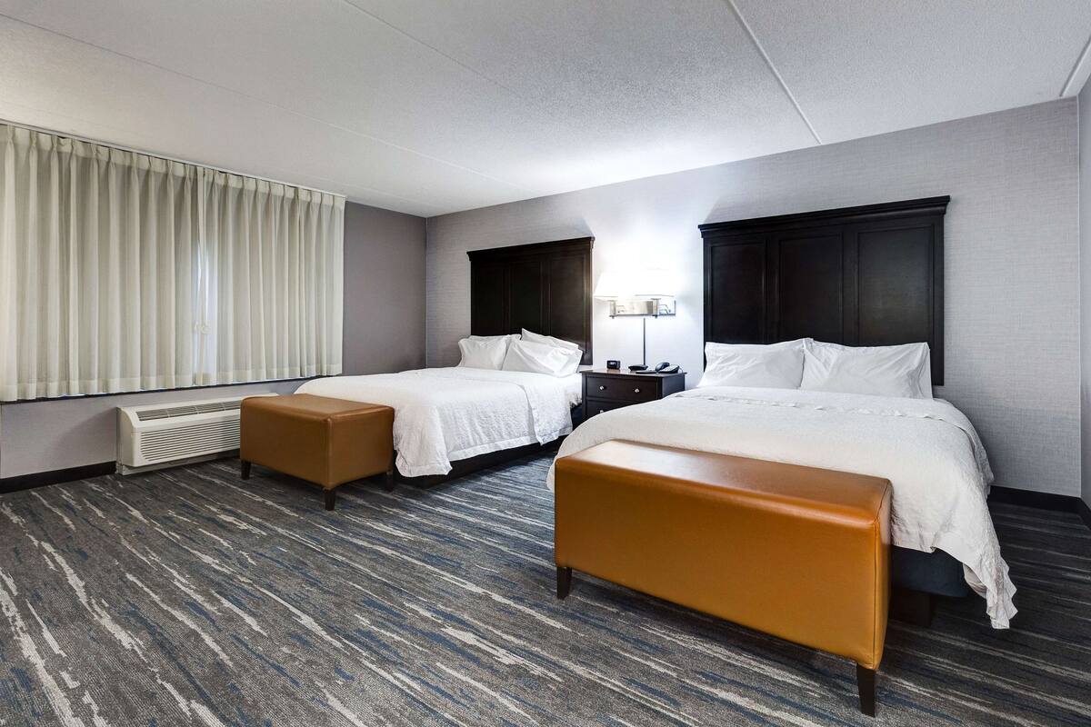 A hotel room with two beds, a large window with curtains, a standing heater, and a carpeted floor.