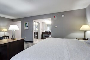 A hotel room with a large bed, two bedside tables with lamps, a desk, a chair, a bathroom visible through a glass door, and a small painting on the wall.