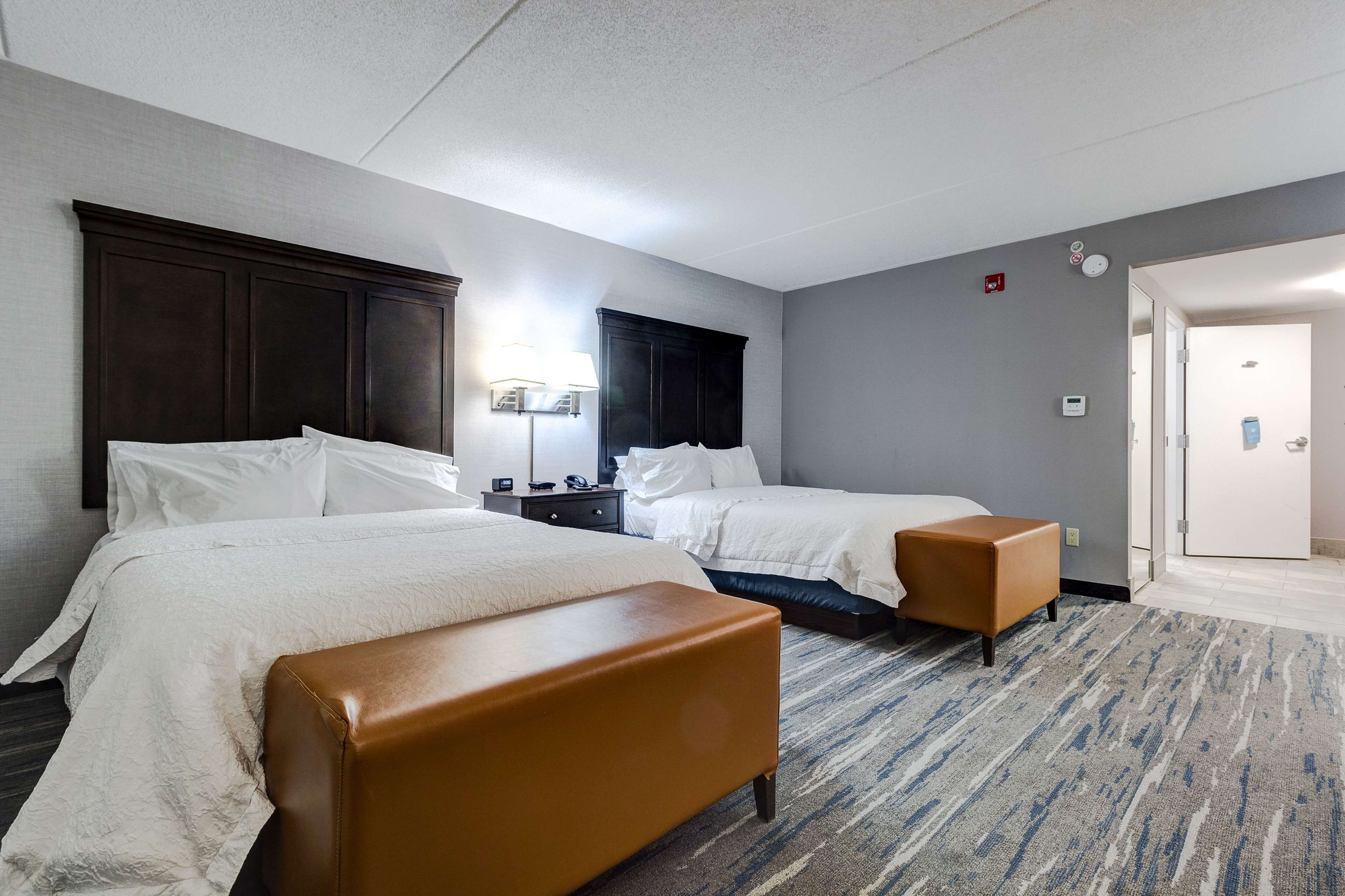 Habitación de hotel con dos camas, un banco de cuero marrón, una mesita de noche con lámpara y una puerta con interruptor de luz.