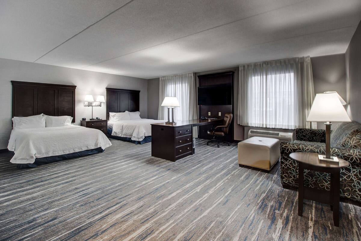 A hotel room with two beds, a desk, a chair, a dresser, and a small table with a lamp.