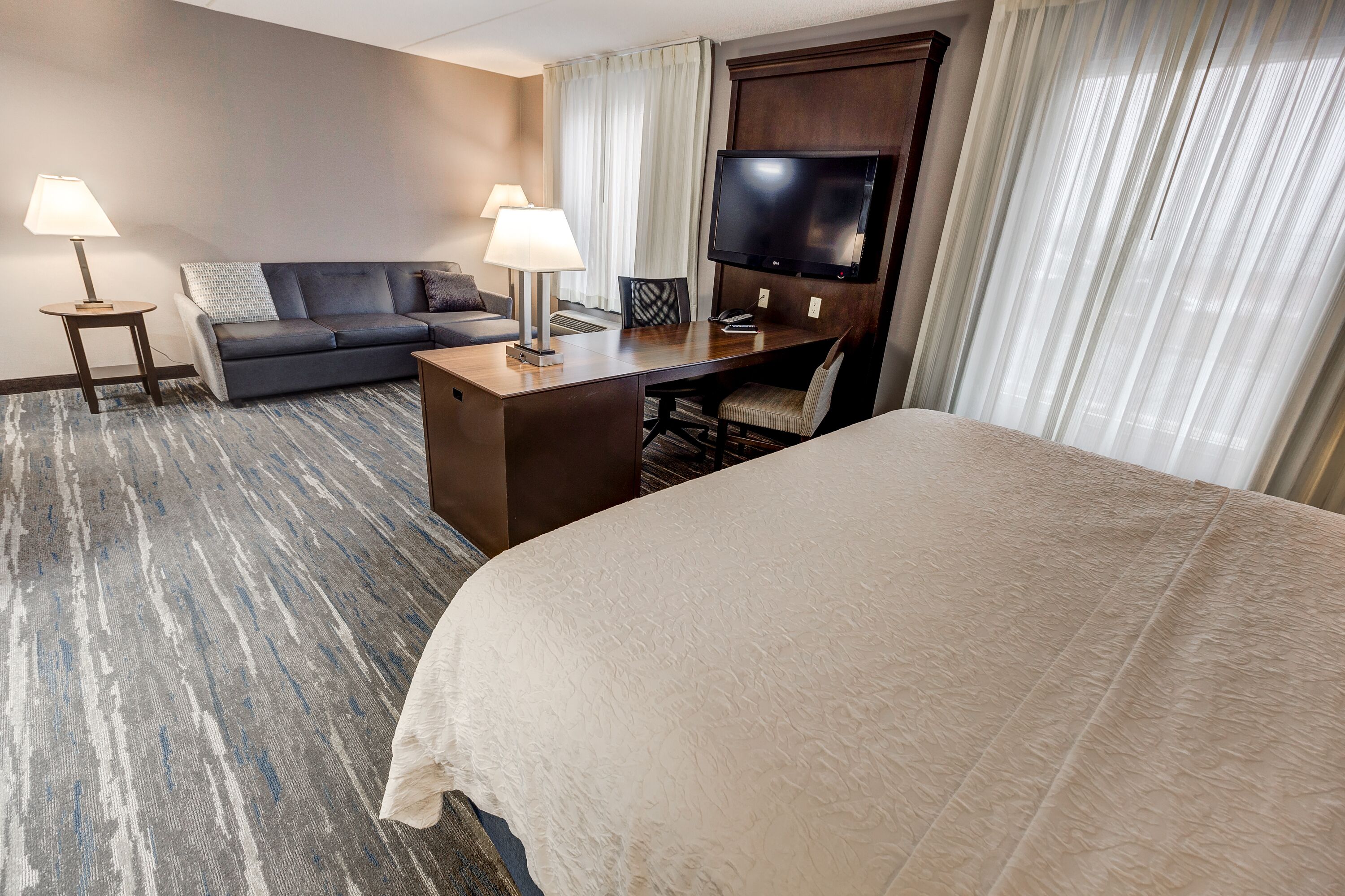 Habitación de hotel con una cama grande, un escritorio con silla, un sofá, dos lámparas, un televisor de pantalla plana y un ventanal con cortinas.