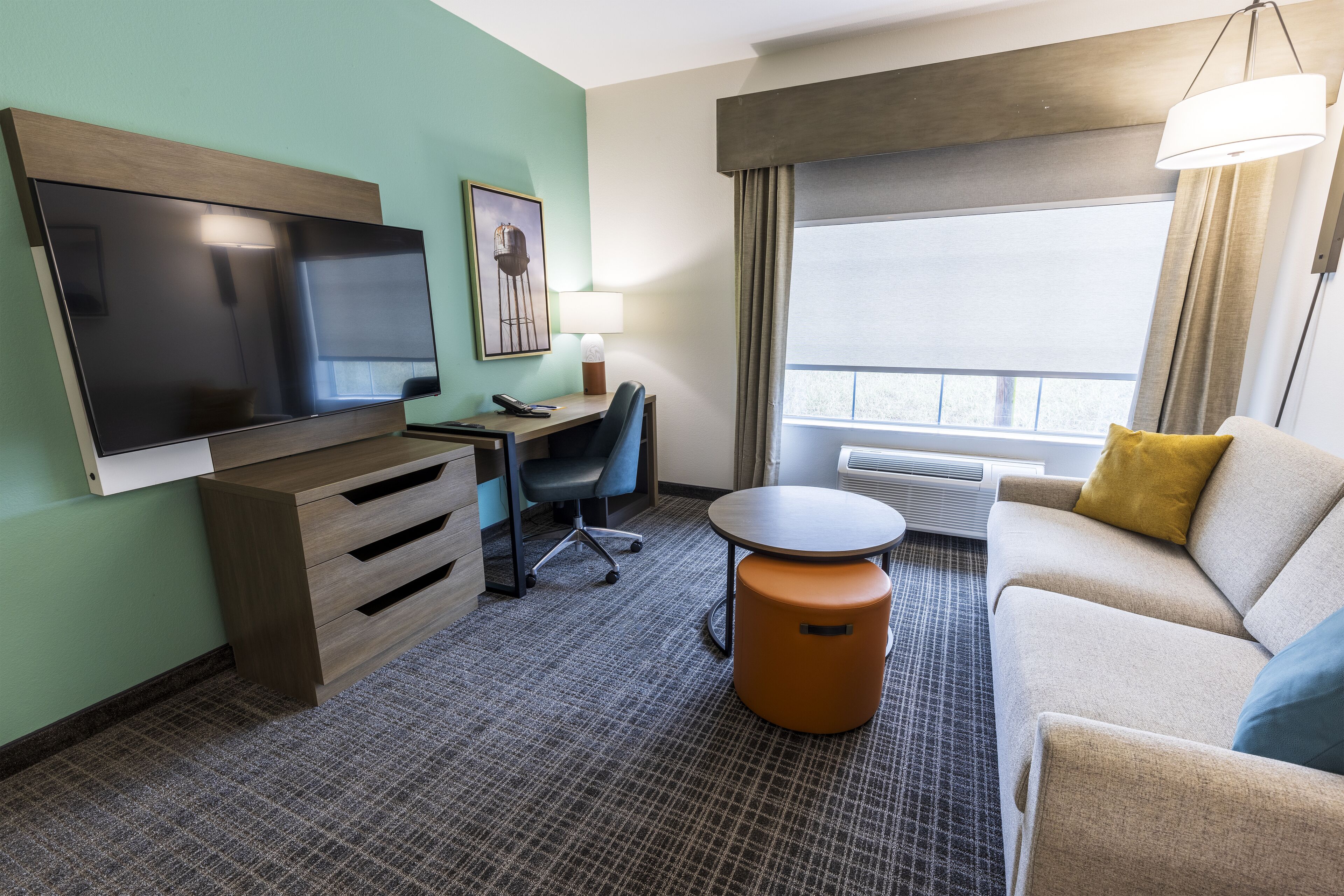 Ein Hotelzimmer mit Flachbildfernseher, Schreibtisch, Stuhl, Sofa, rundem Hocker und einem großen Fenster mit Vorhängen.