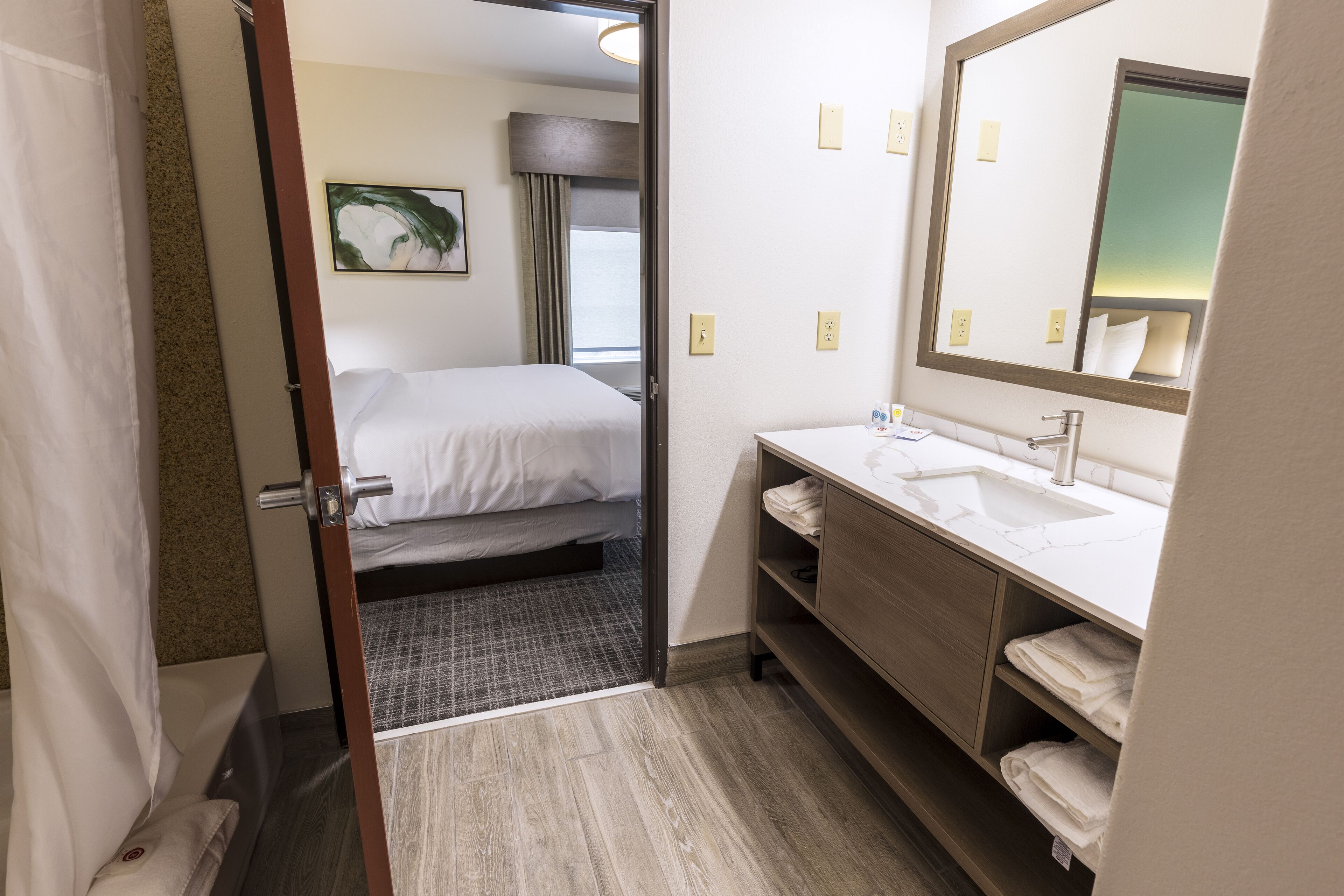 Una camera d'albergo con un letto, un bagno con lavandino e specchio, e una porta che dà su un'altra stanza.