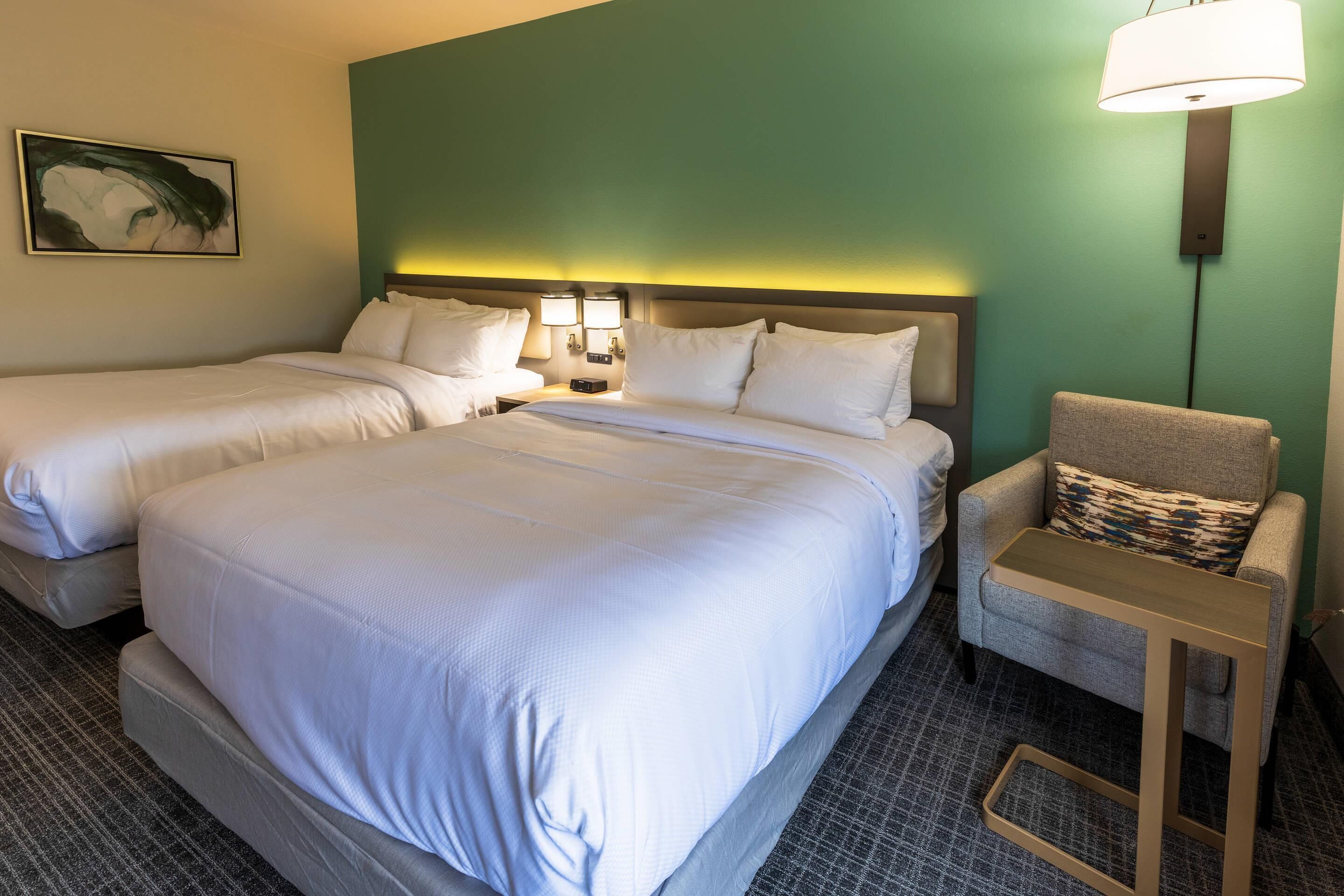 A hotel room with two beds, a nightstand, a chair, and a wall-mounted lamp.