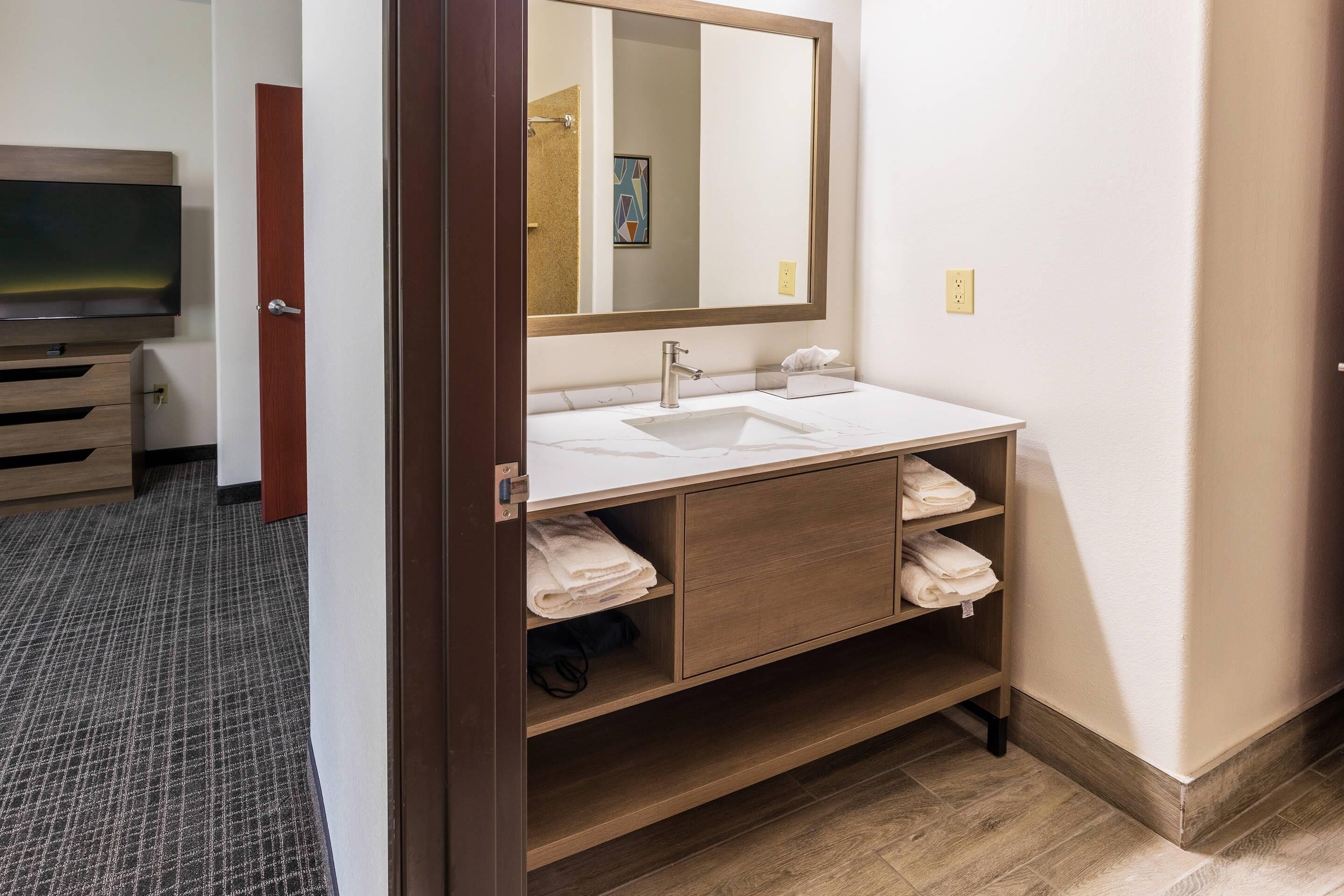 A hotel bathroom with a large mirror, a sink, a wooden vanity, and a television in the adjacent room.