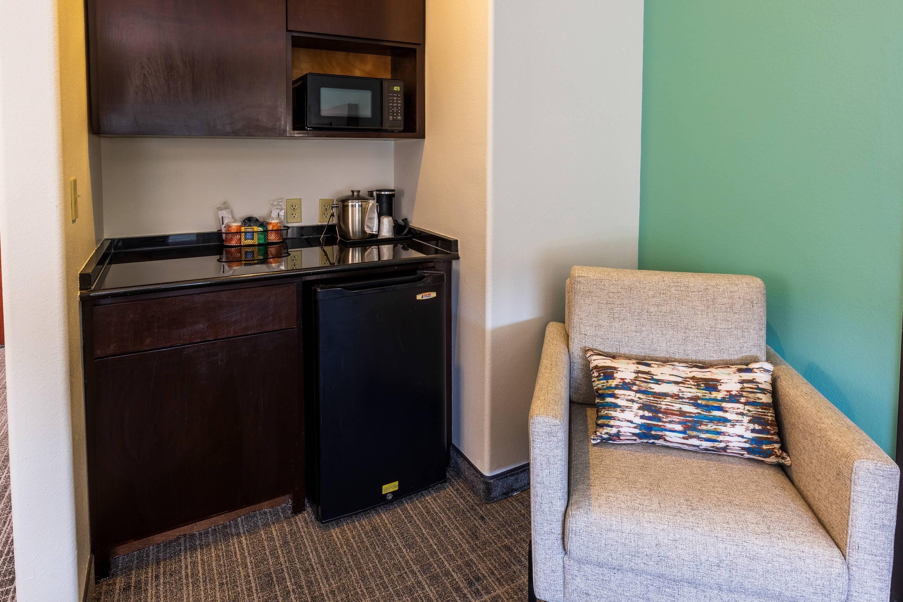 Una kitchenette compacta con microondas, cafetera y refrigerador pequeño. Una silla con un cojín estampado está ubicada frente a una pared verde.