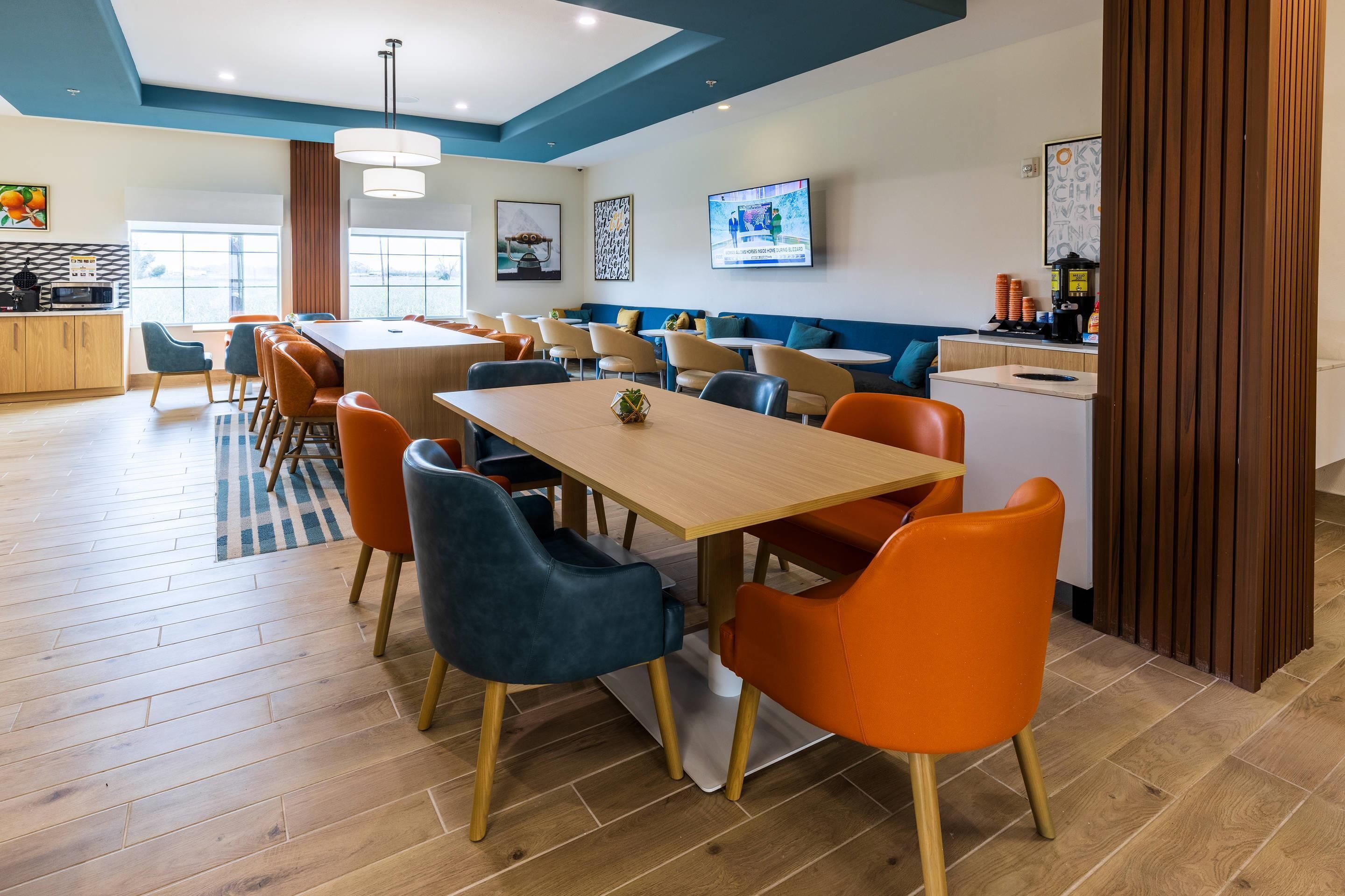 A modern hotel dining area with multiple tables and chairs, a kitchen in the background, and artwork on the walls.