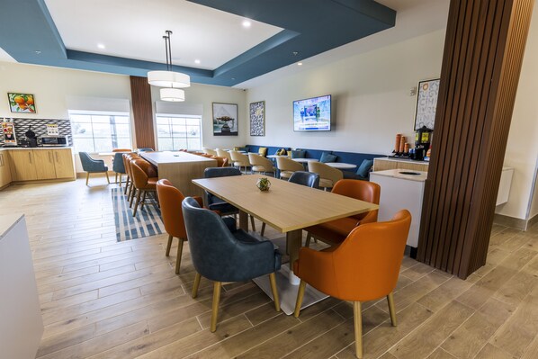 Un espace repas moderne avec une longue table, des chaises orange et bleues, et une cuisine en arrière-plan.