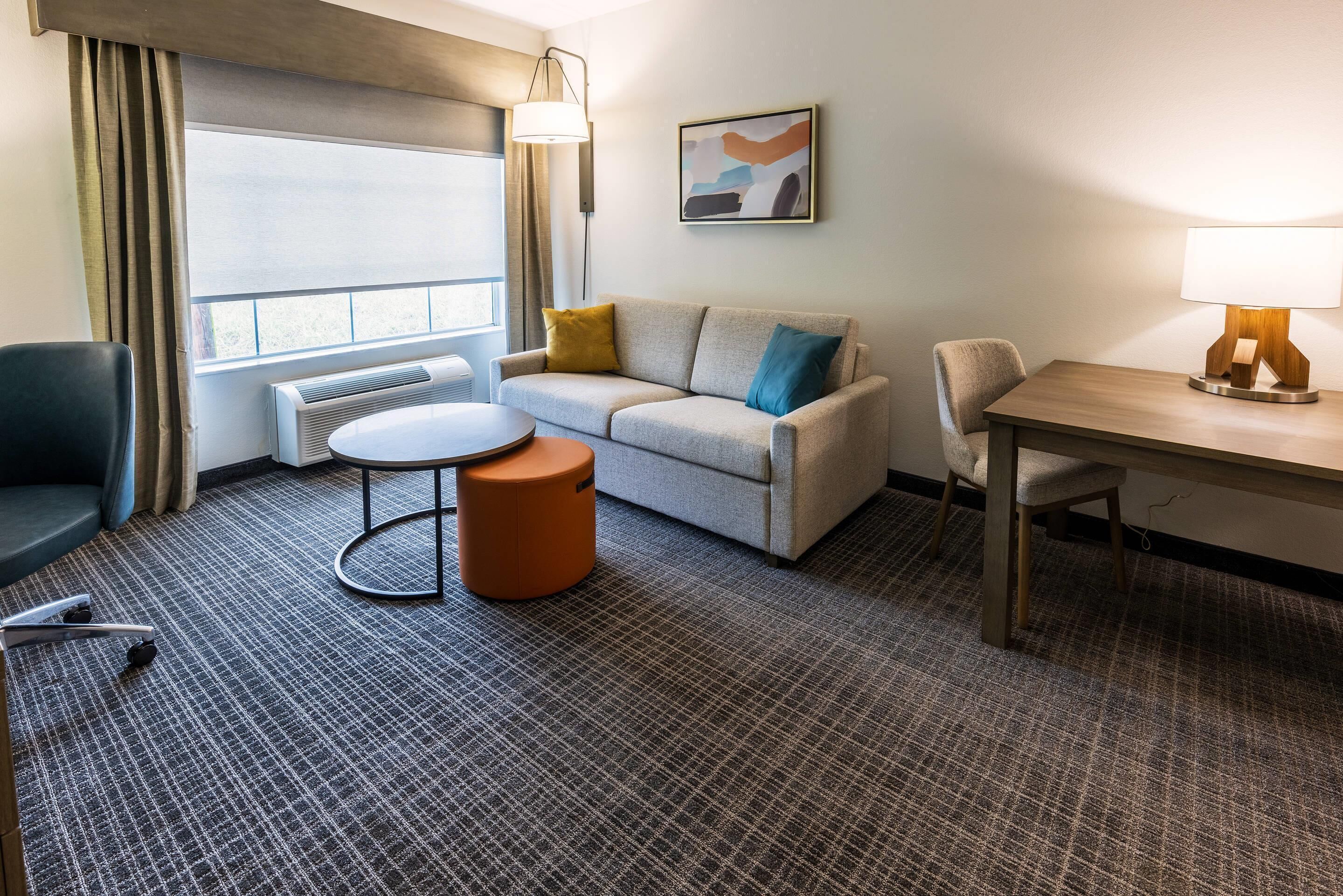 A hotel room with a sofa, a small round table, a chair, a lamp, and a painting on the wall.