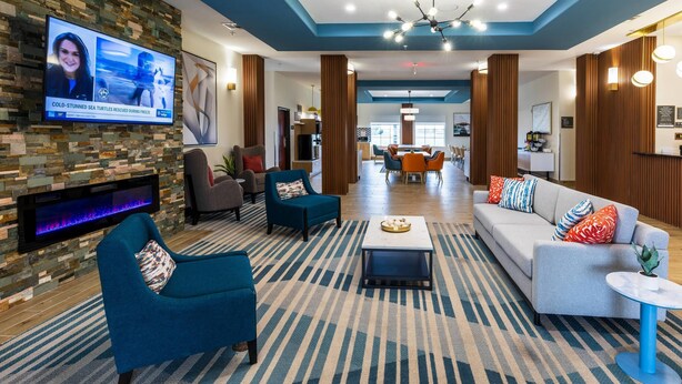 A hotel lobby with a TV displaying news, a fireplace, seating areas, and a dining area.