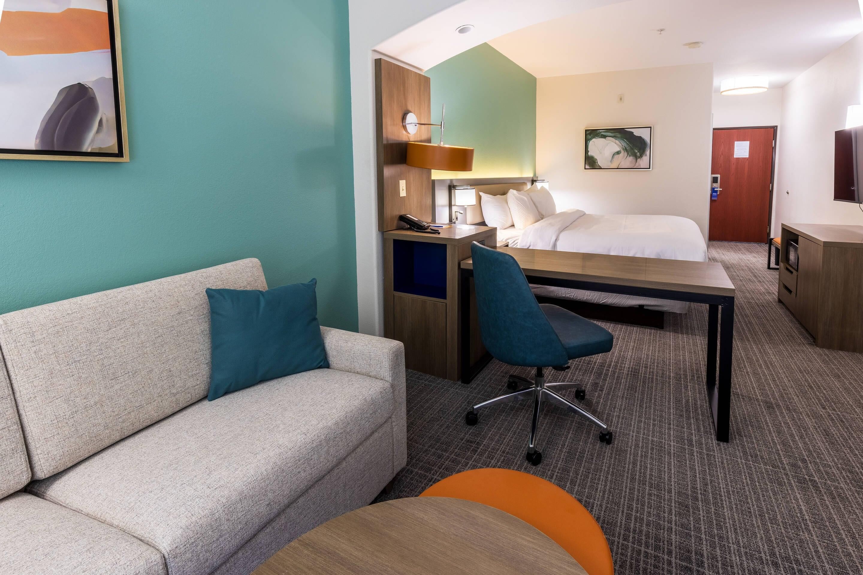 A hotel room with a sofa, a desk, a chair, a bed, a TV, and a painting on the wall.