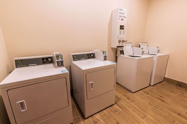 A laundry area with three front-loading machines, a wall-mounted soap and bleach dispenser, and a wooden floor.
